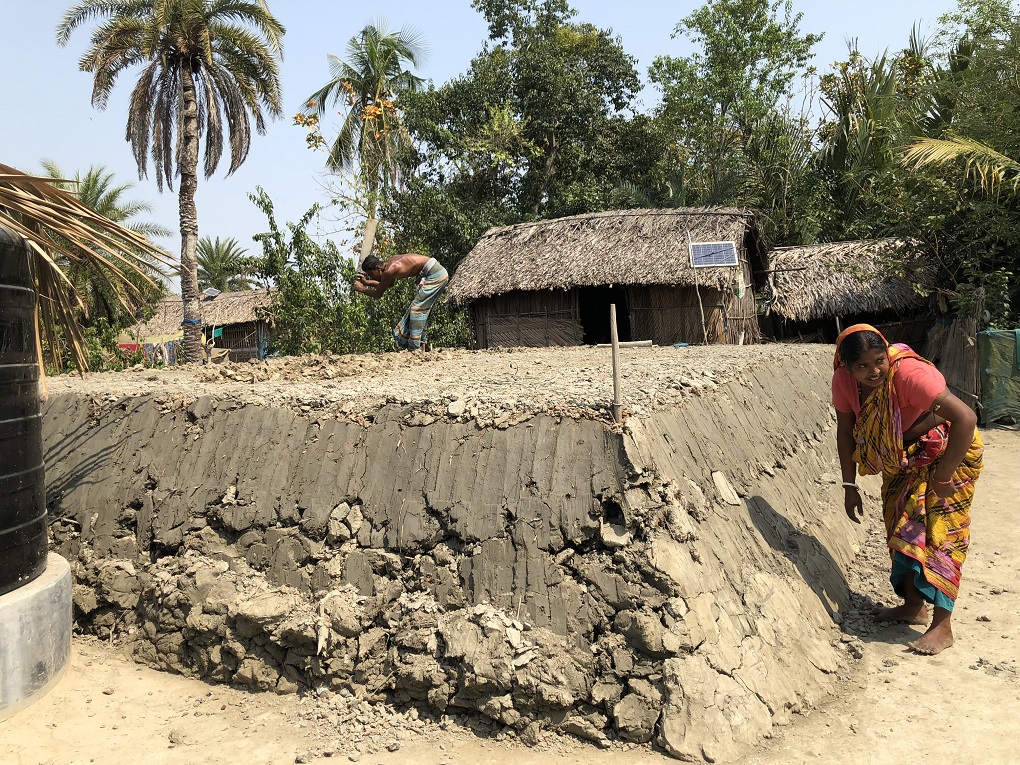 <p>In Joymonigola village near the port of Mongla in southern Bangladesh, Tripti Mandal shores up the four-foot-high mud platform that husband Kishore is completing, so that they can raise their hut (in the background), hopefully above the high tide level this monsoon [image by: Joydeep Gupta]</p>