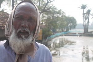 <p>In a village on the edge of the Sundarbans and facing the Bay of Bengal, Jasimuddin Sarkar in front of his farm that now grows nothing, thanks to sea level rise, and his house that gets flooded with dirty saline water every third or fourth day [image by: Joydeep Gupta]</p>