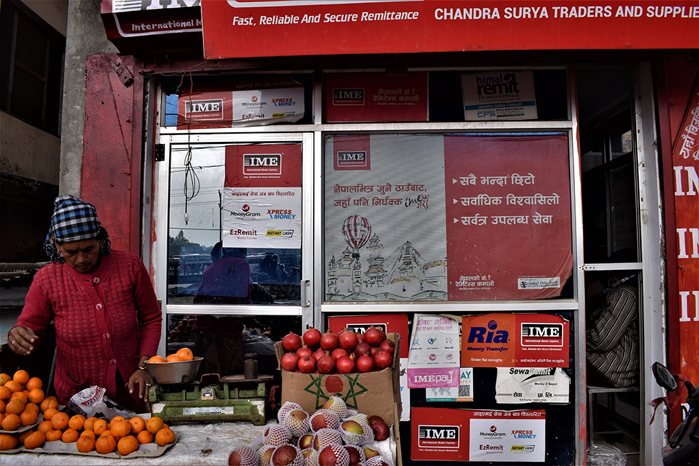 A woman runs a fruit stall in front of an advertisement for a remittance delivery firm along banks of Mahakali