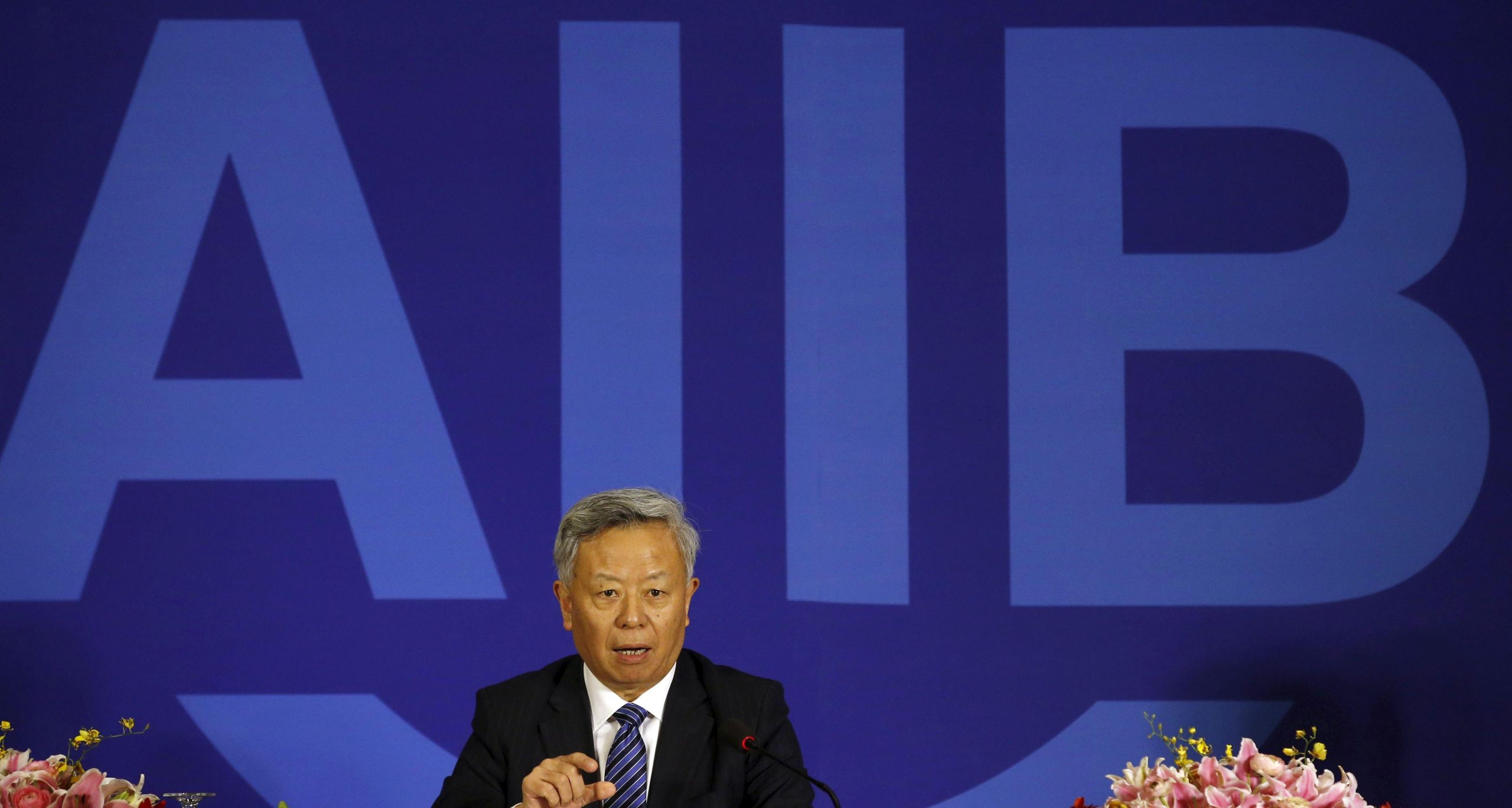 A man in a suit and tie sitting in front of a sign that reads AIIB