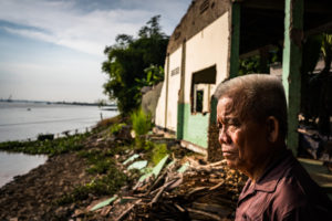 Nguyen Van Thuong, 71 stands in front of his former neighbors house which has been destroyed and fallen into the river as a result of erosion