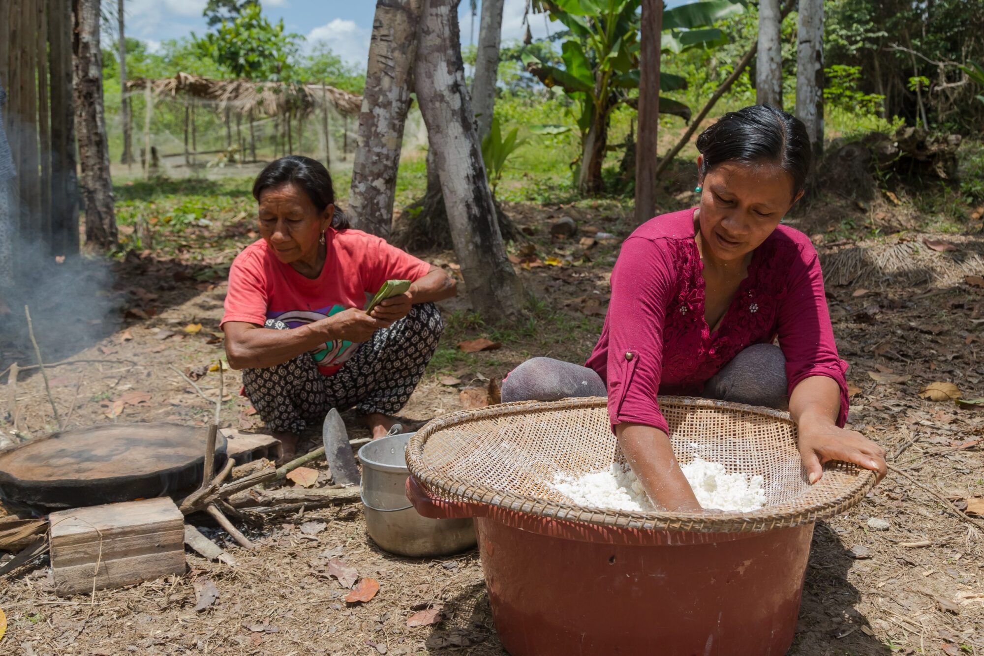 Liz Chicaje Churay crouching with a woman in the background, filtering cassava. 