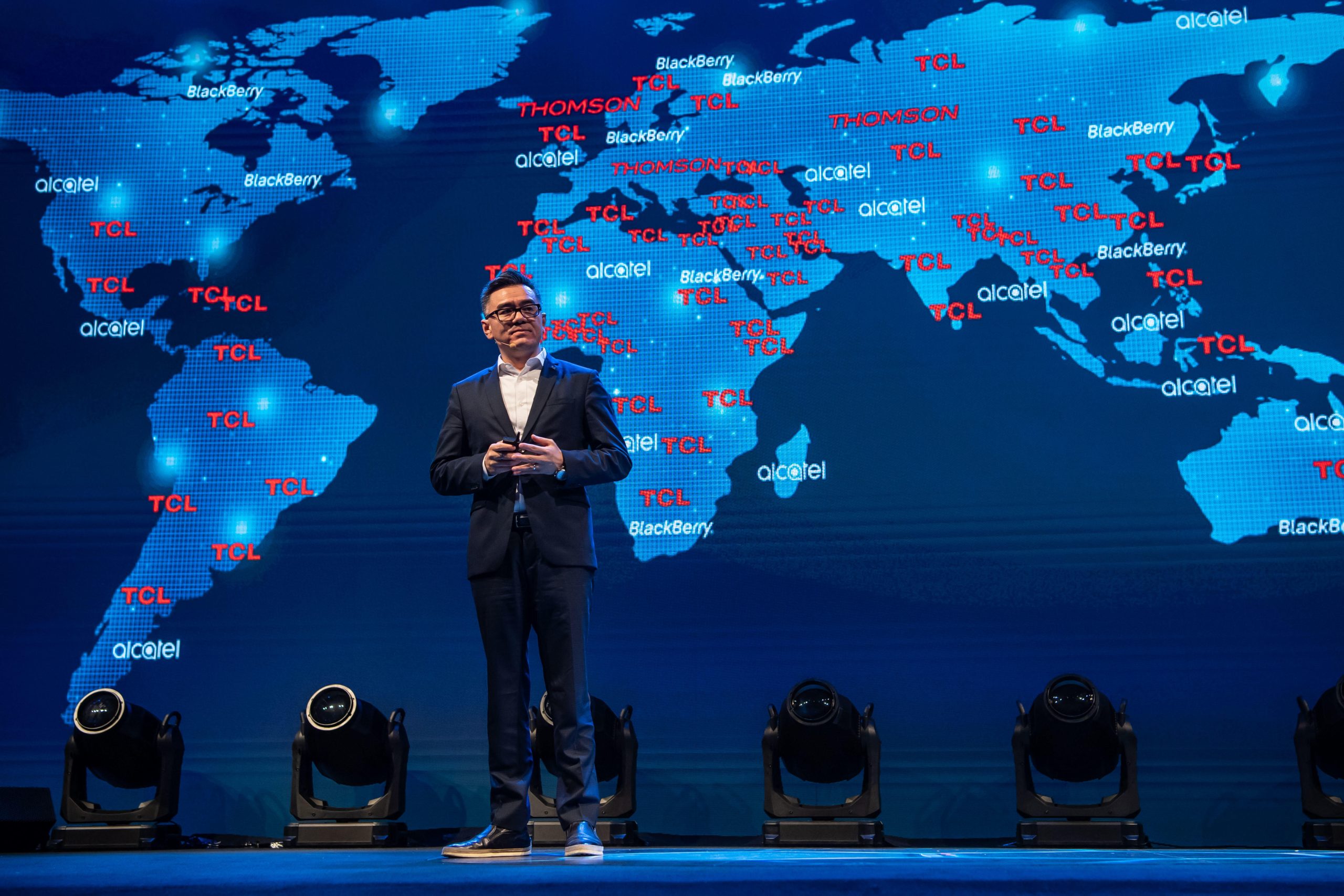 A man in a suit standing on a stage with a map of the world behind him.