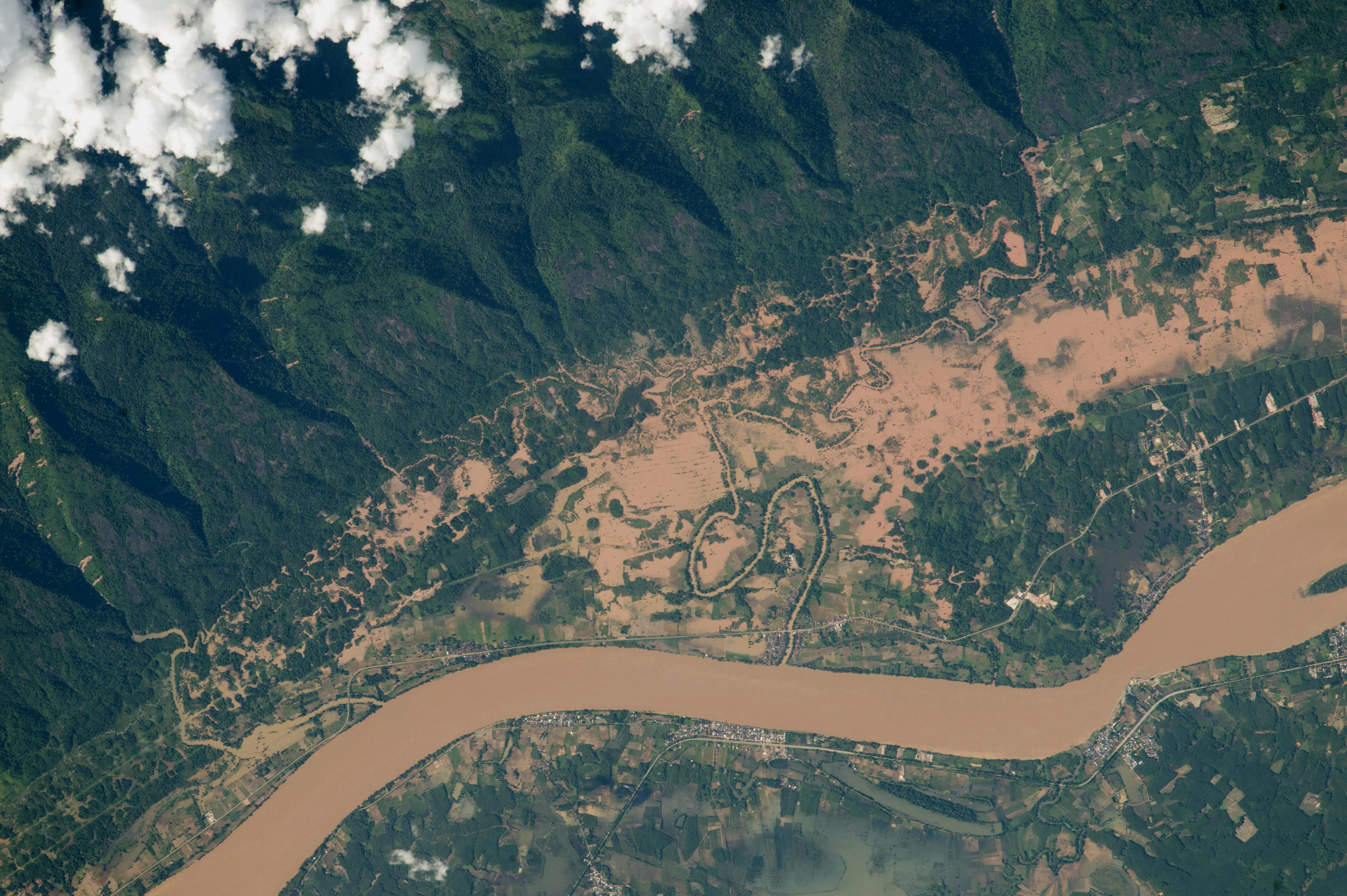 Above cloud overhead view of brown river with flooded regions along the bank bordered by green mountains