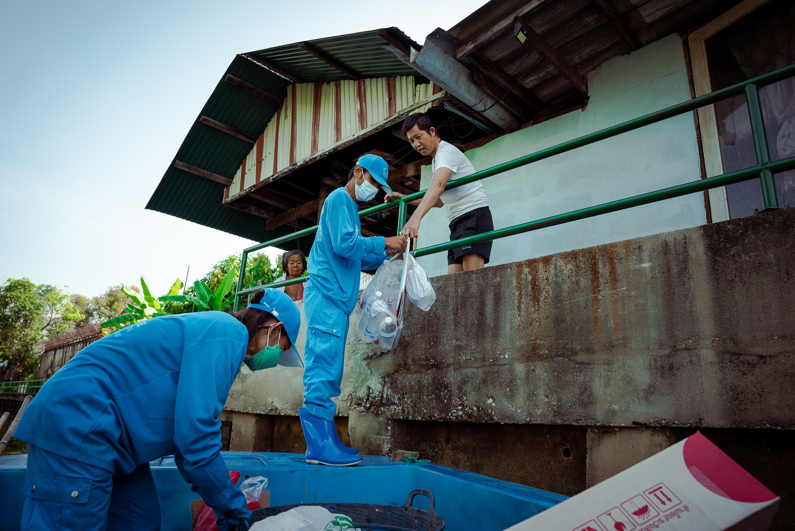 man on balcony handing plastic waste to men in blue uniforms