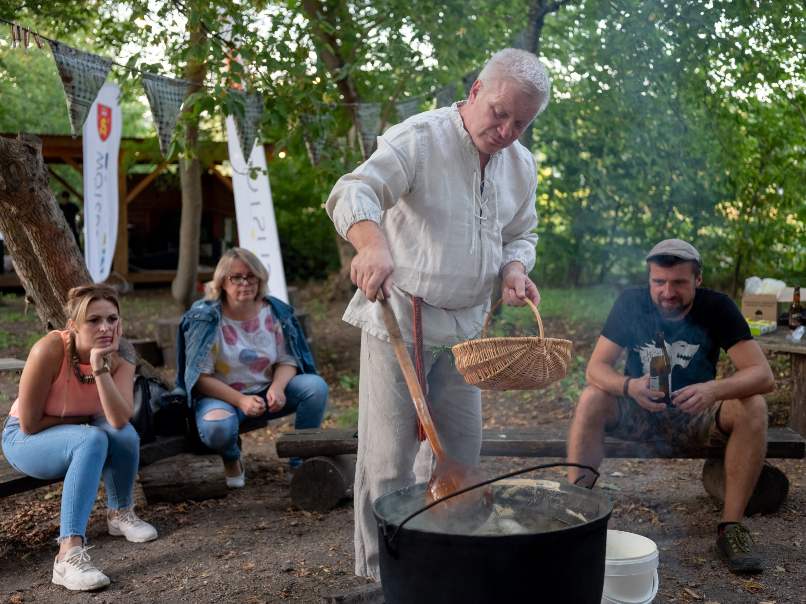 A man in white traditional clothing stirs a pot of brewing mead over a fire in the forest