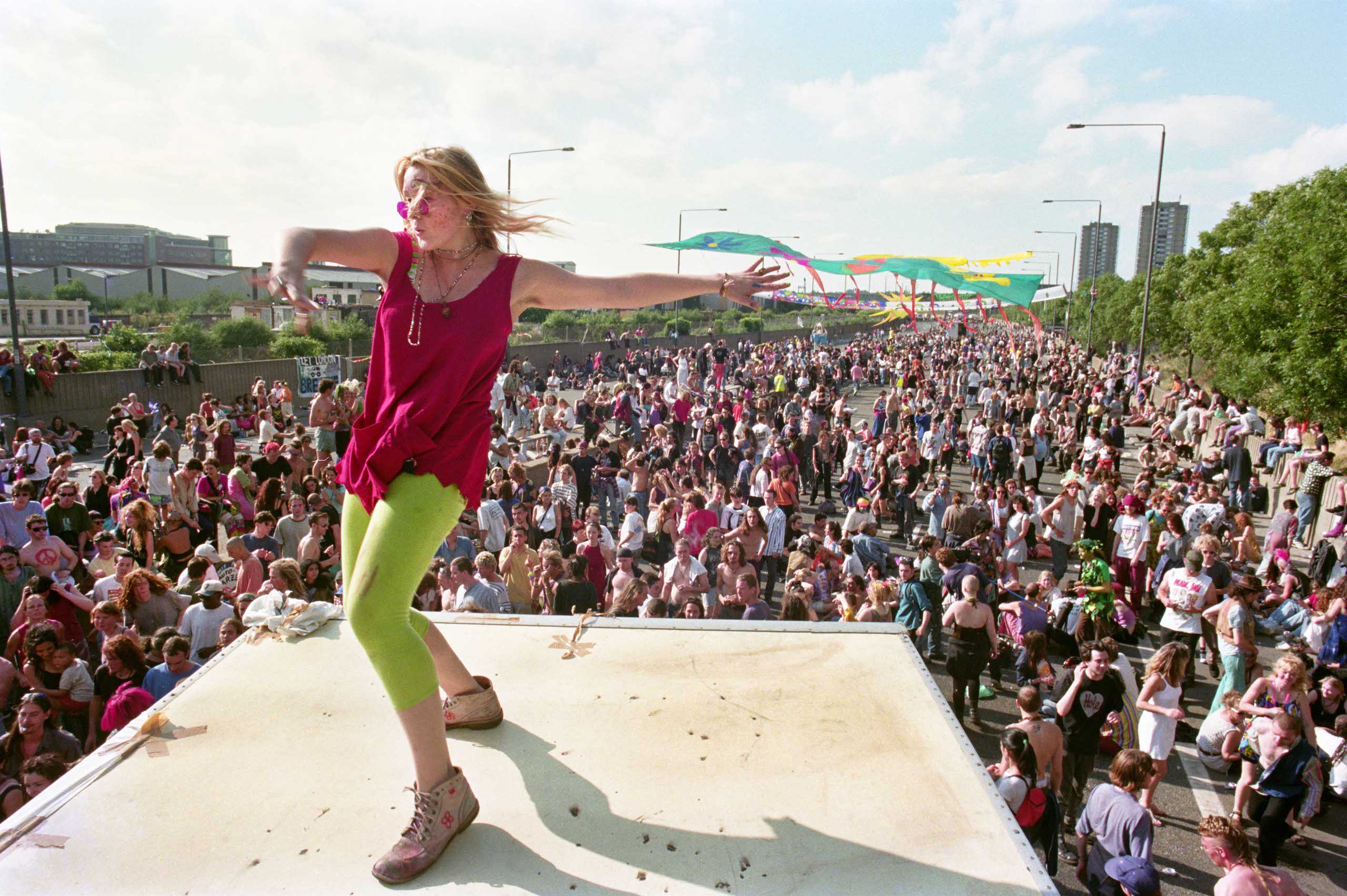 A woman dances atop a lorry as a crowd dances on the motorway behind. Historical photograph