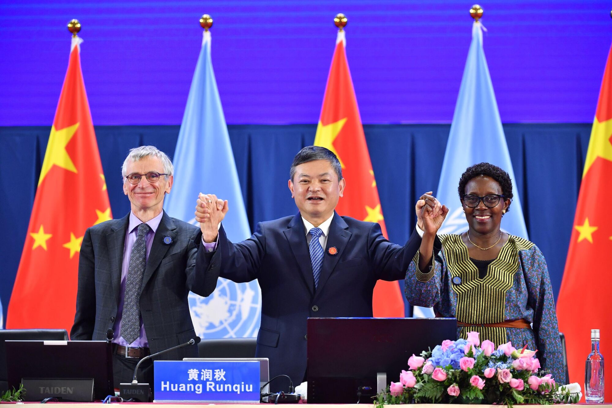 Dois homens e uma mulher de mãos dadas, em frente às bandeiras da ONU e da China.