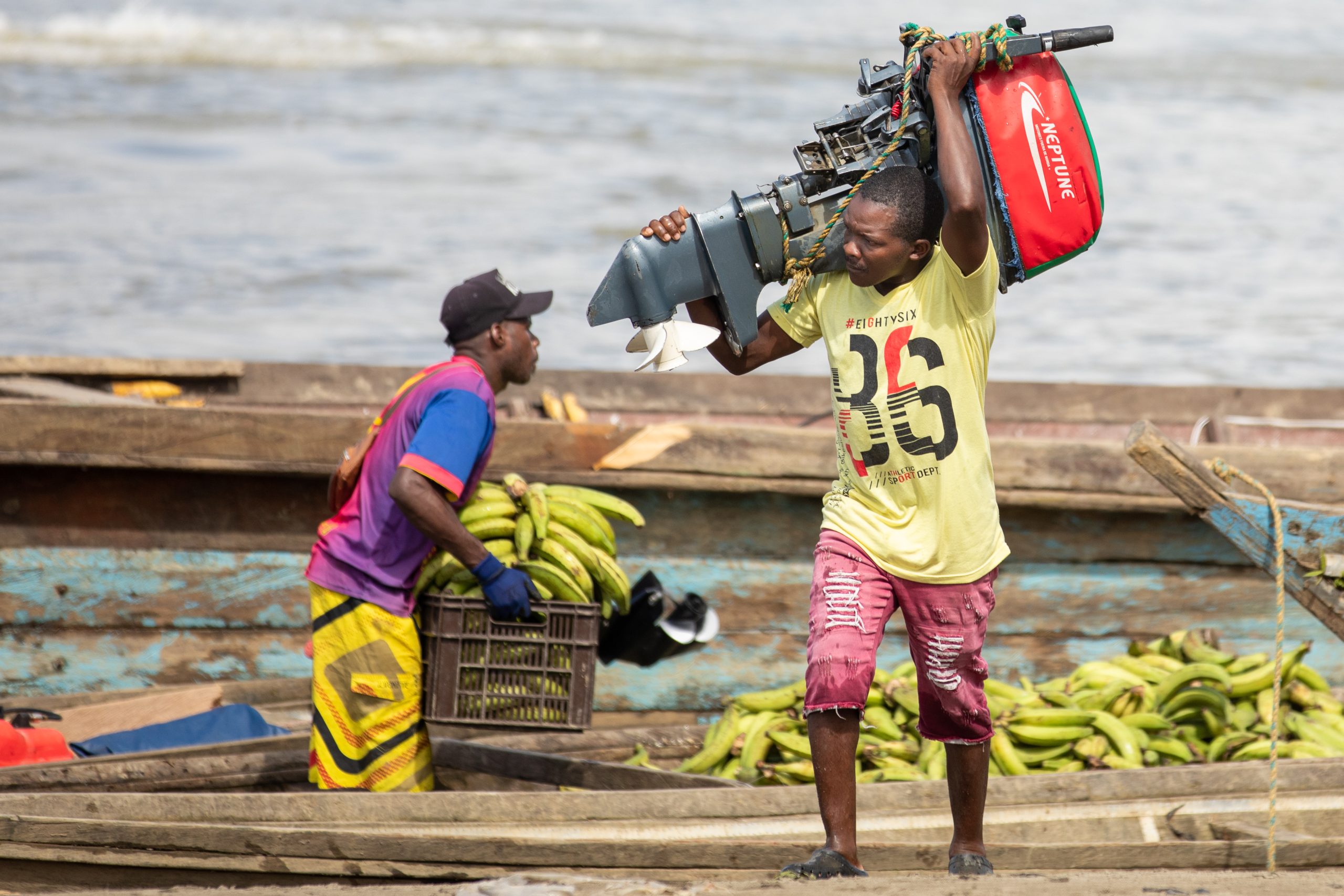 one man offloading crate of plantains while second carries motor on small boat