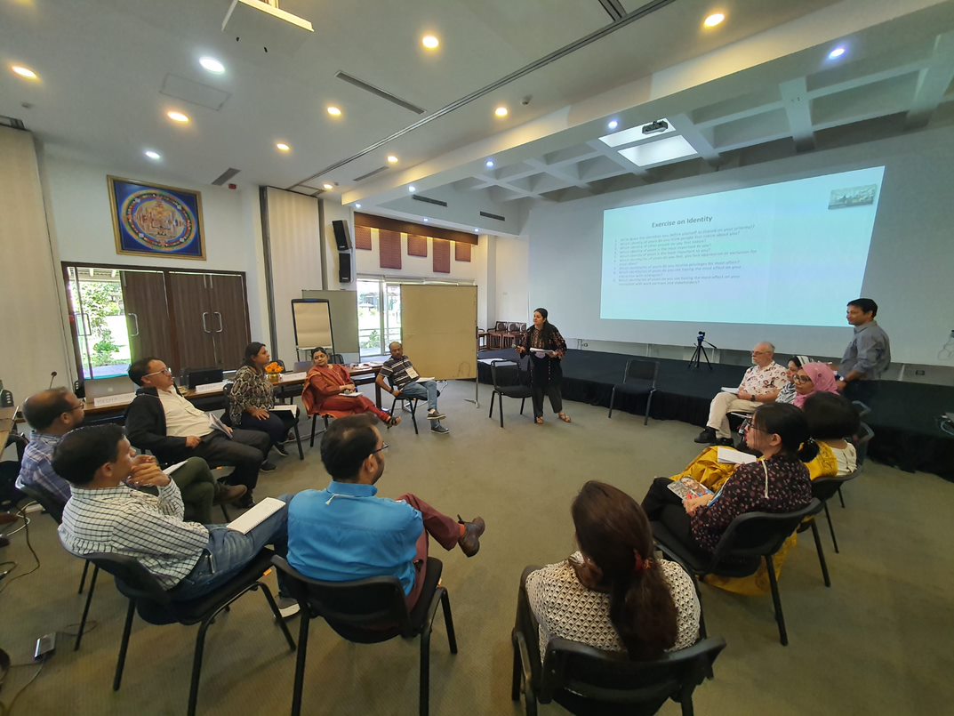 Adults sat in a circle of chairs around a woman standing giving a lecture in front of a projector screen