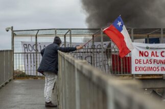 Un hombre que protesta por el cierre de una planta de fundición de cobre ondea la bandera de Chile ante humo, asomado a un balcón.