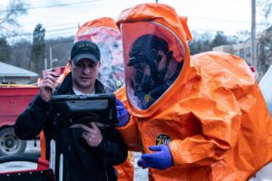 Members of Ohio National Guard, one wearing an orange hazmat suit and the other pointing at a tablet computer, prepare to enter the area affected by the East Palestine chemical spill