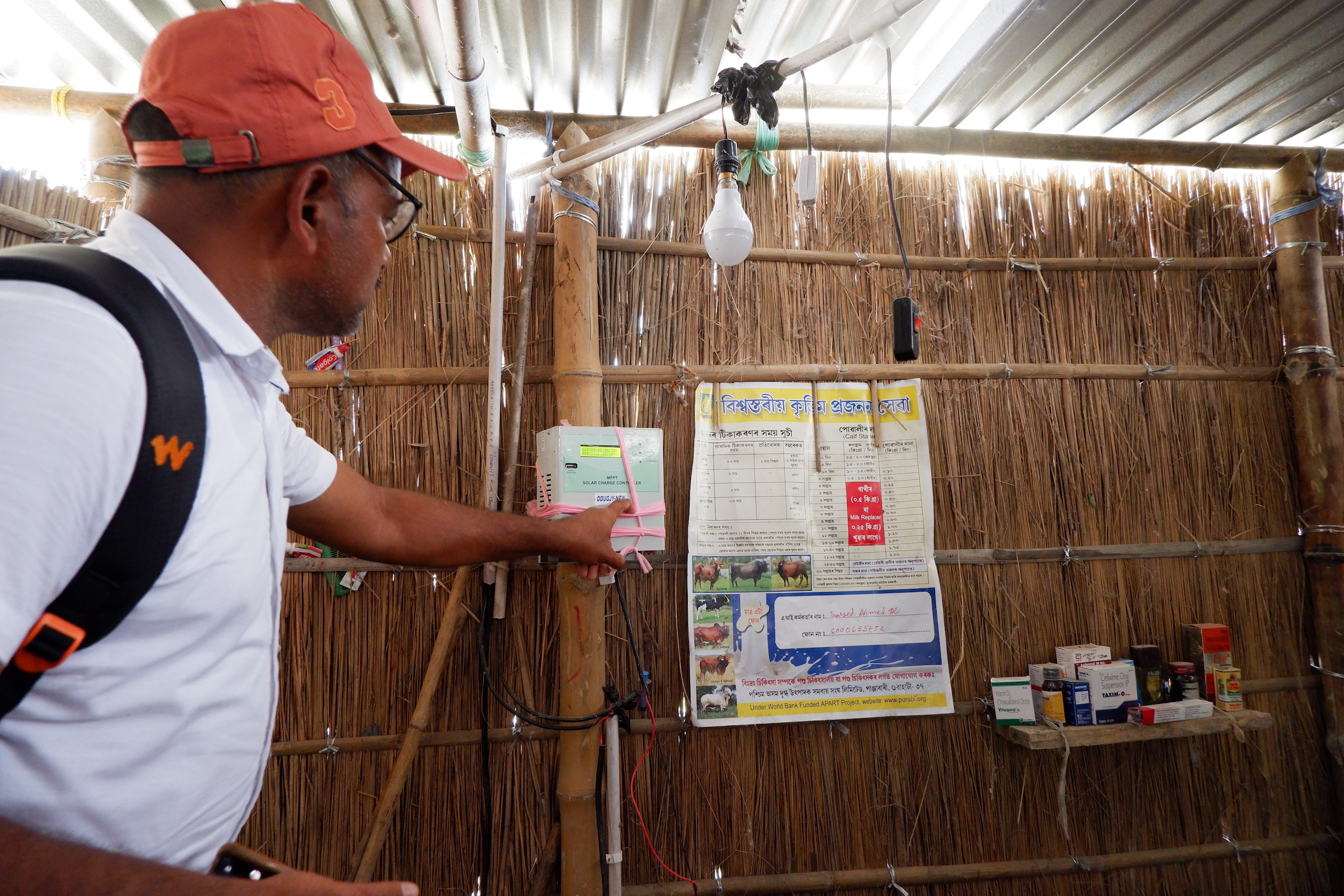 Man in white polo shirt and orange cap, Abdul Kalam, gestures toward a solar charging system provided under a government scheme, Assam