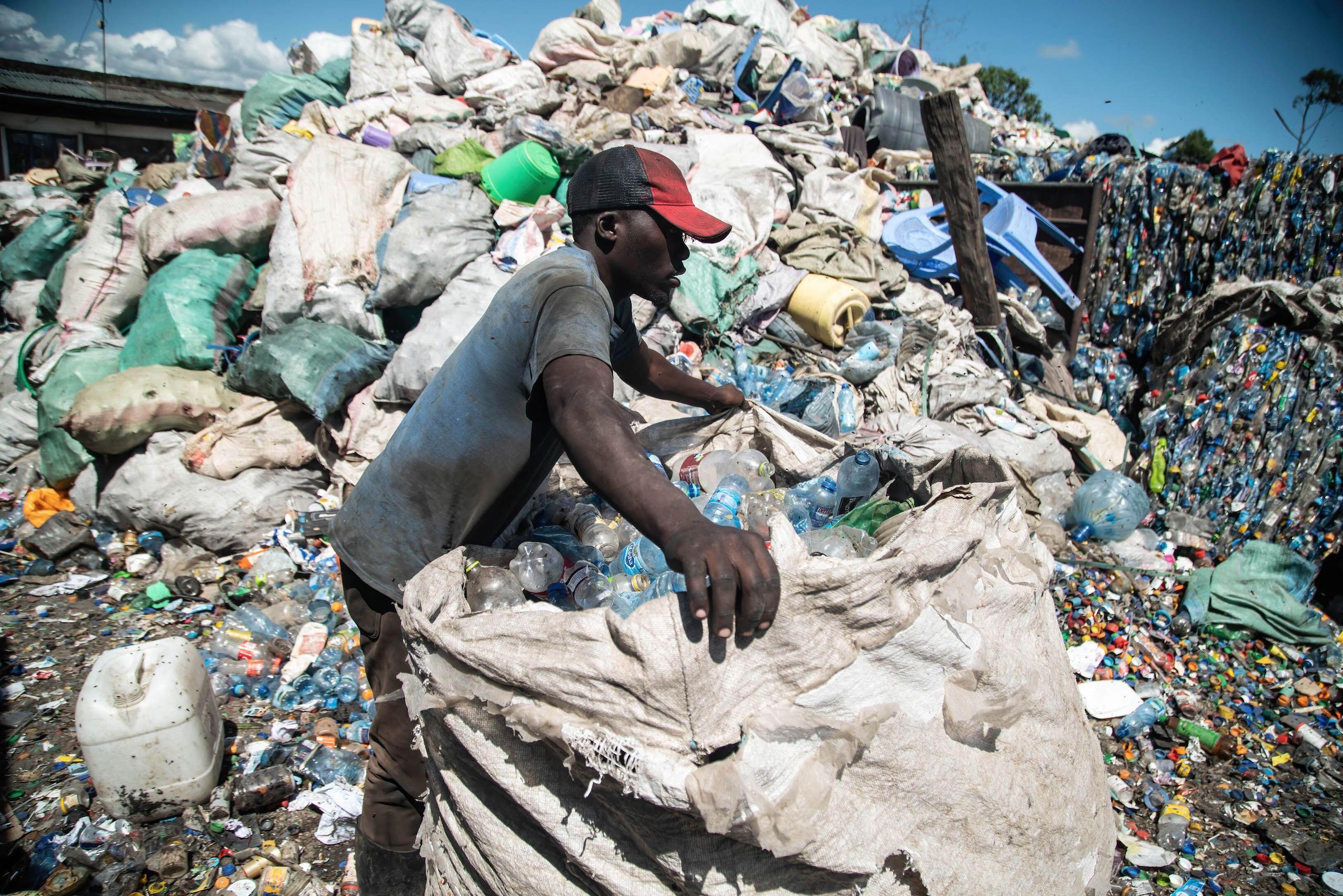 Man wearing a red cap lifts a large bag full of waste plastic bottles in front of a huge pile of plastic bottles to be recycled