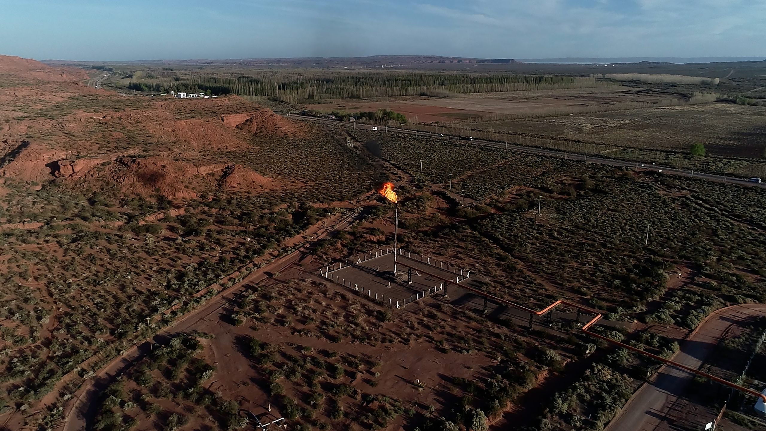 Flames from gas flaring pipe in the centre of a flat scrubland landscape, orange-red soil and forest in the distance