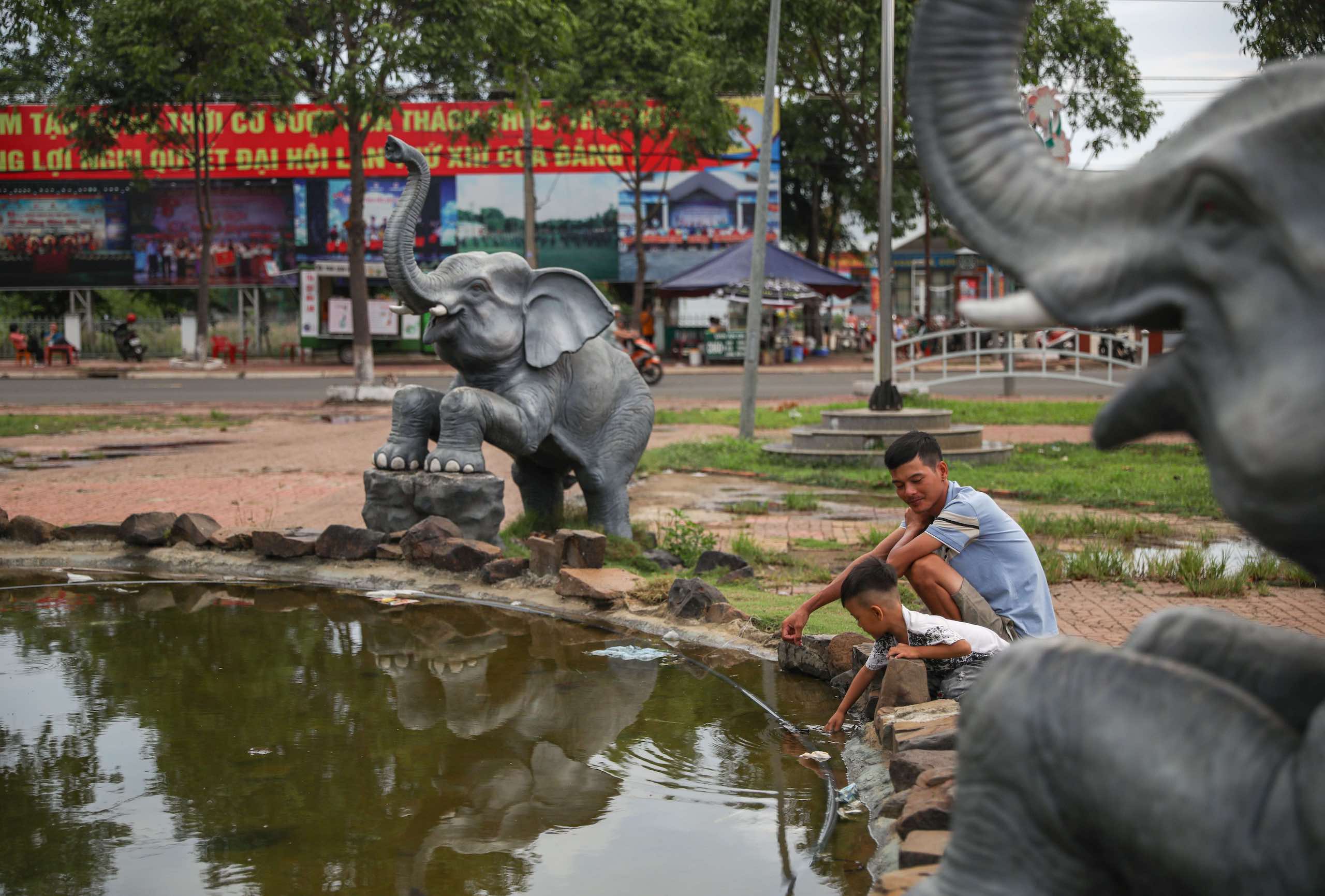 A fruit vendor playing with his 3-year-old son at a fountain surrounded by elephant statues