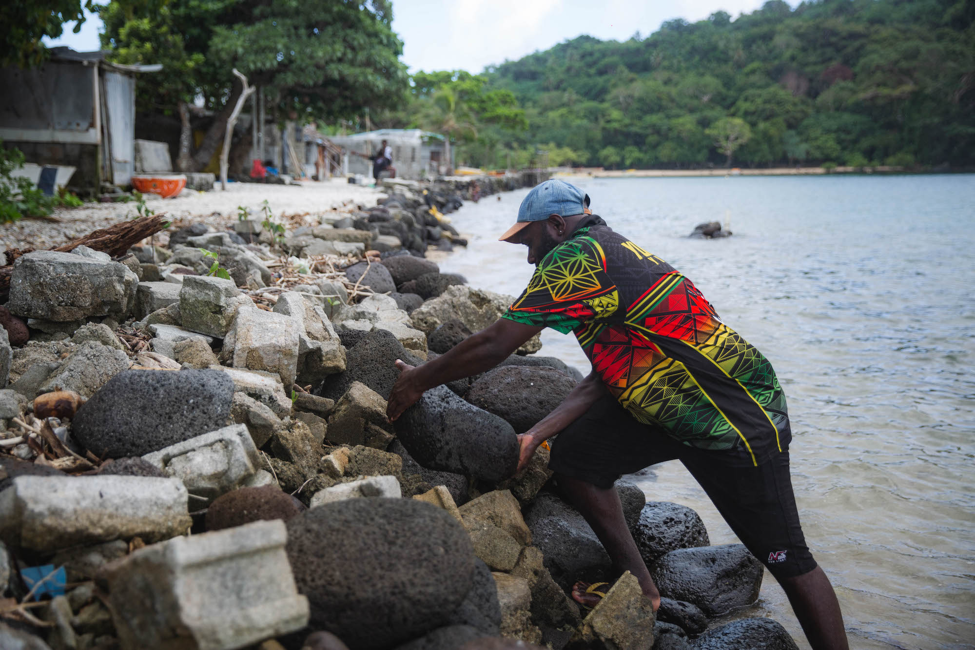 Man in colourful shirt places a stone in a seawall, coastal forest in background