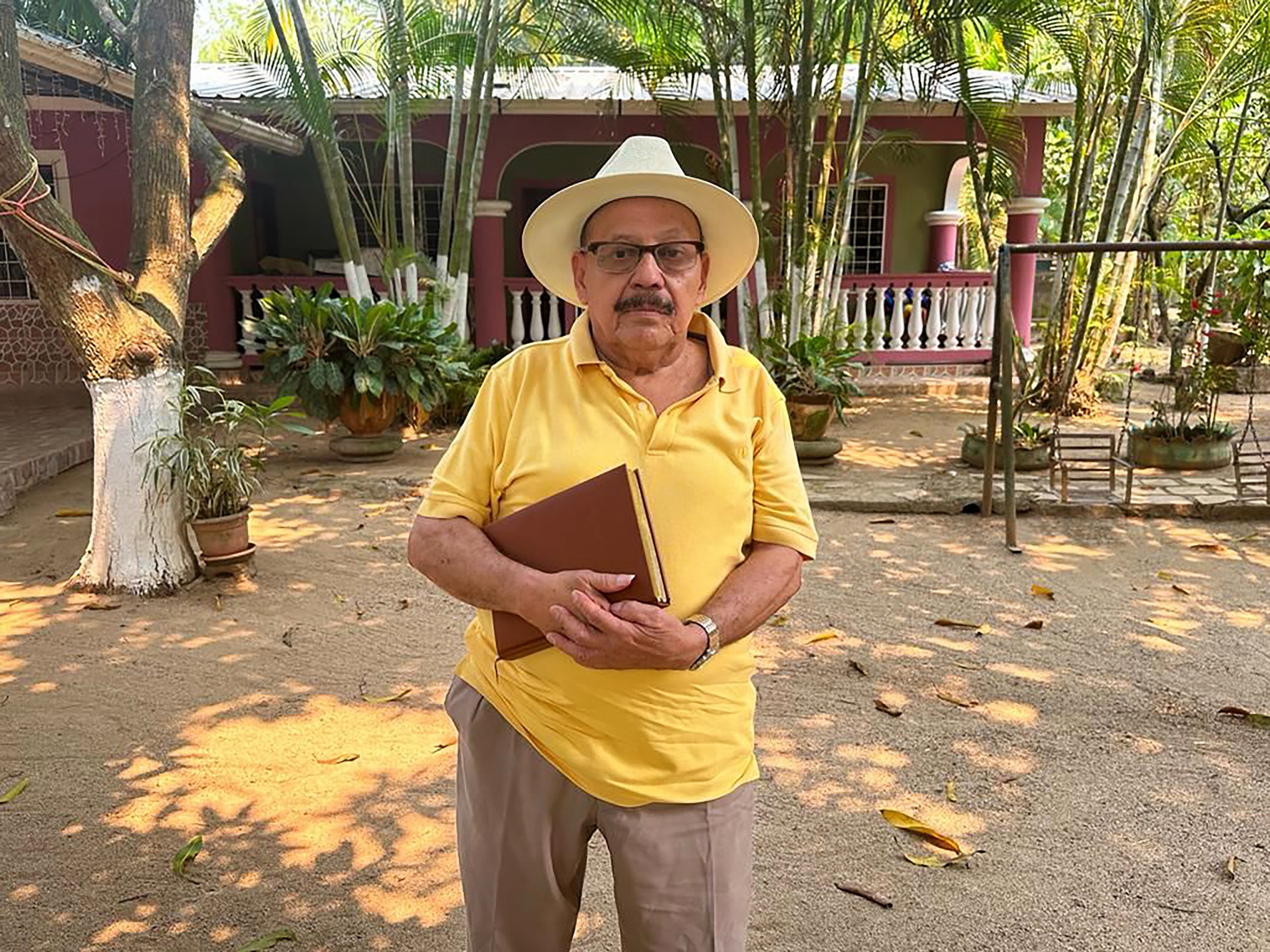 Un hombre con una remera amarilla y sombrero blanco sostiene un libro, en un patio