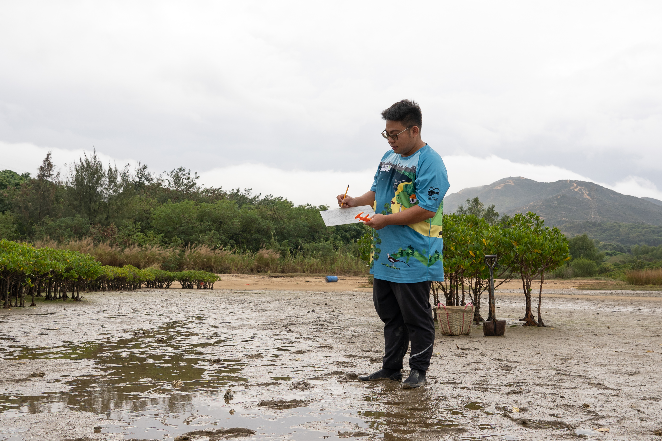 A man takes notes on a clipboard standing on a beach by mangrove trees