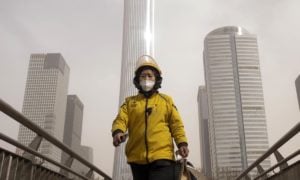 Person wearing a face mask and yellow jacket walks in front of tall grey skyscrapers