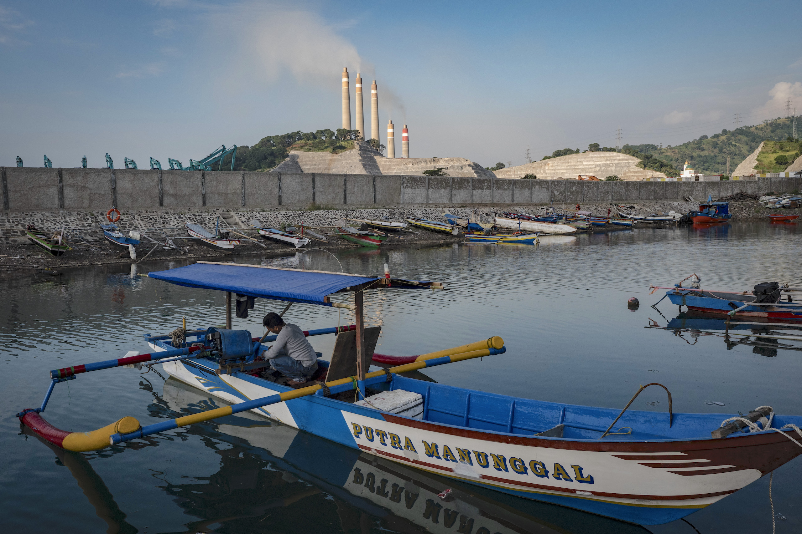 A fisherman prepares his boat at a port with smokestacks in the background