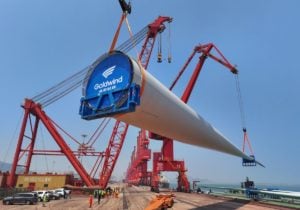Large red crane lifts a wind turbine blade, with the word 'Goldwind' printed on the end, onto a ship at a port