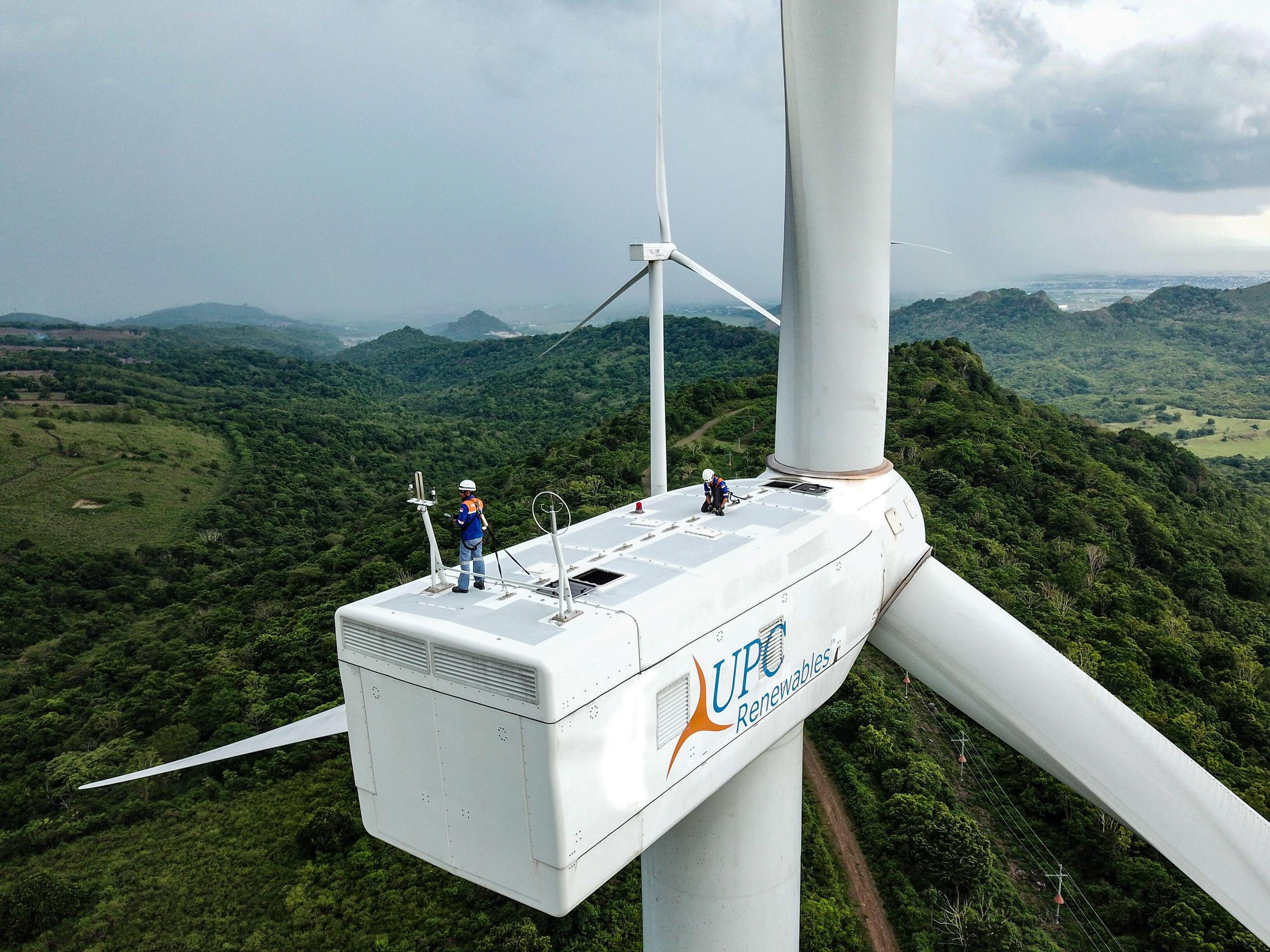 Aerial photo of a two people standing on top of a wind turbine in a wooded mountainous area