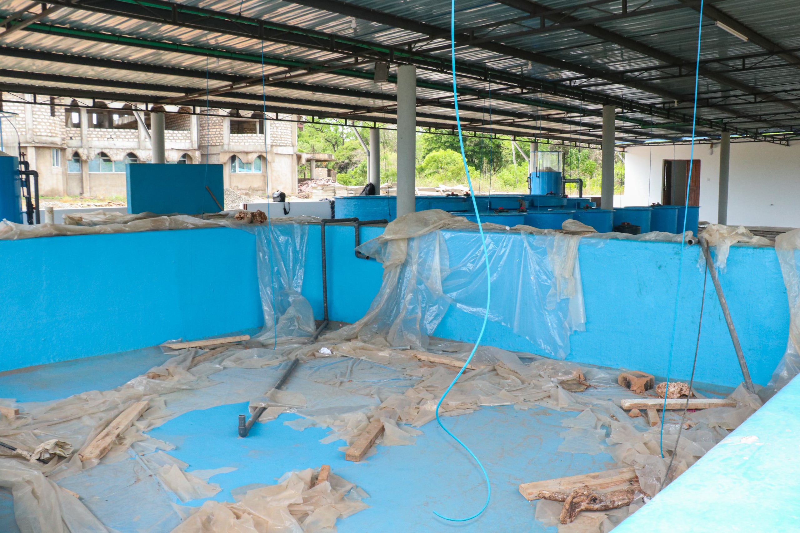 Inside of a building without walls, containing large blue tanks and plastic sheets on the floor