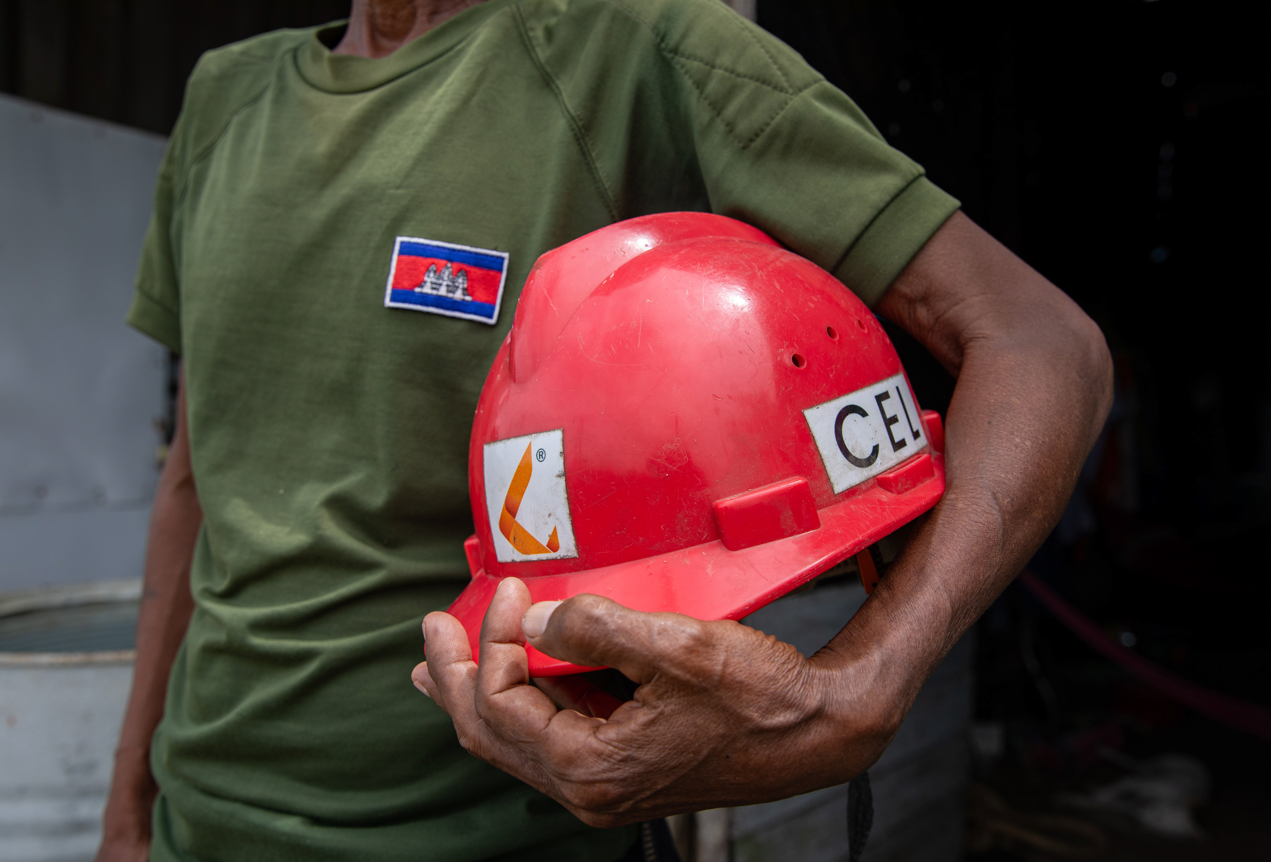 A man, face out of shot, wearing a t-shirt bearing the Cambodia flag holds a red safety helmet under his arm