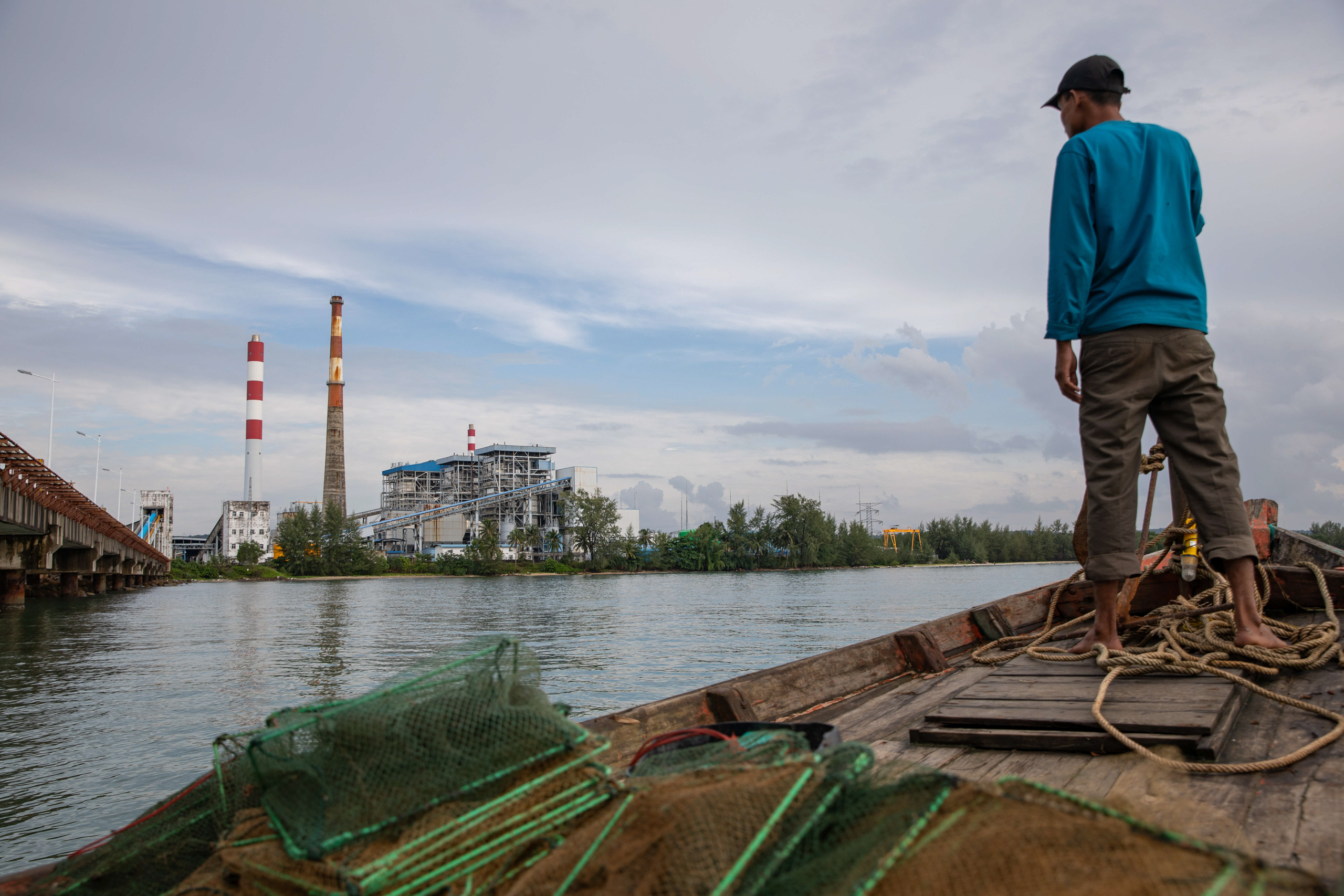 Man stands on a boat looking at an industrial building with smokestacks across the water