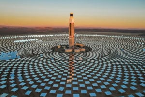 A tower in the centre of a solar power plant, pattern of shiny panels on the ground at sunset