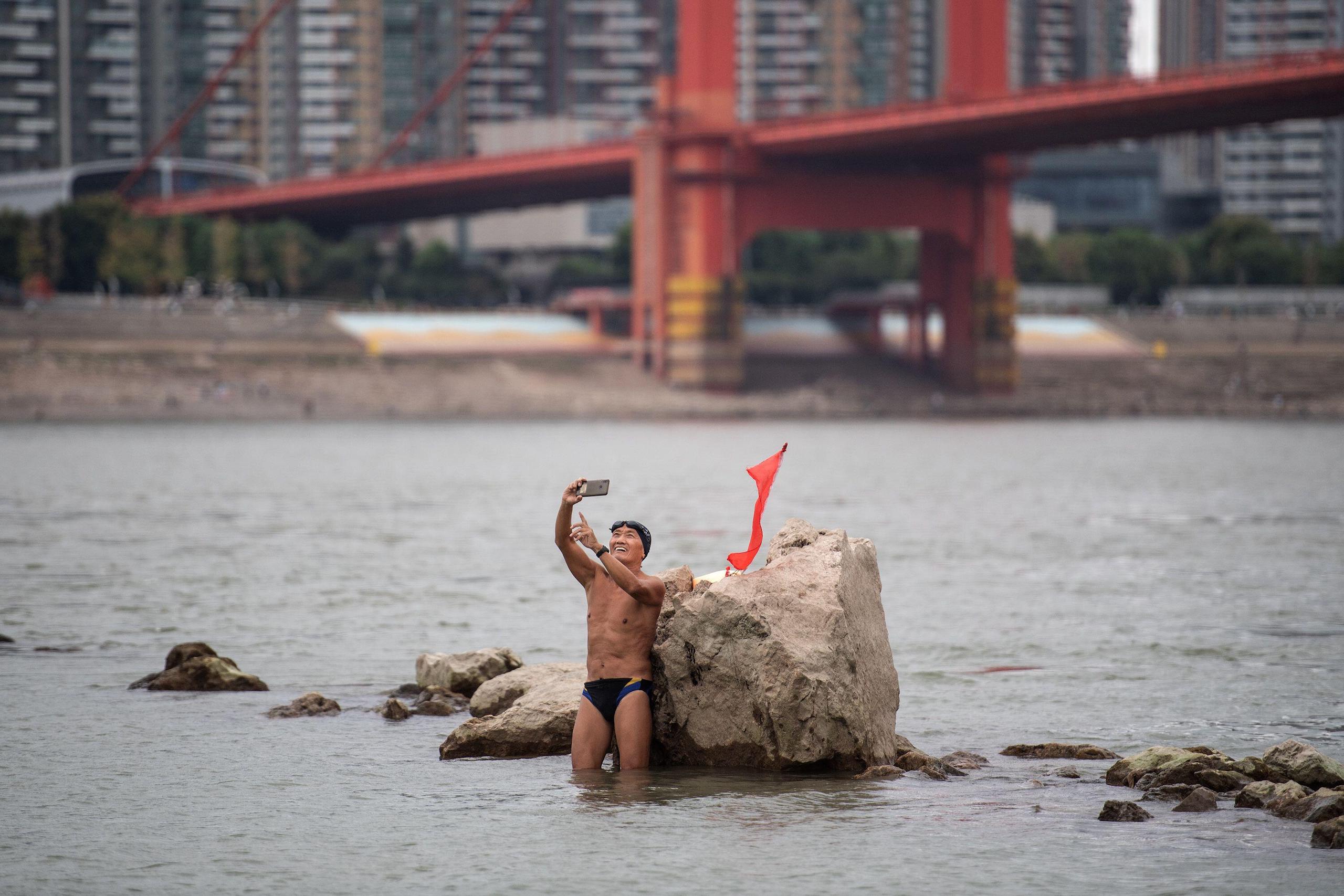 Man in a swimming costume takes a selfie standing in a shallow river