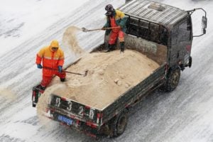 two men in high visibility clothing shovel salts onto icy road from truck bed