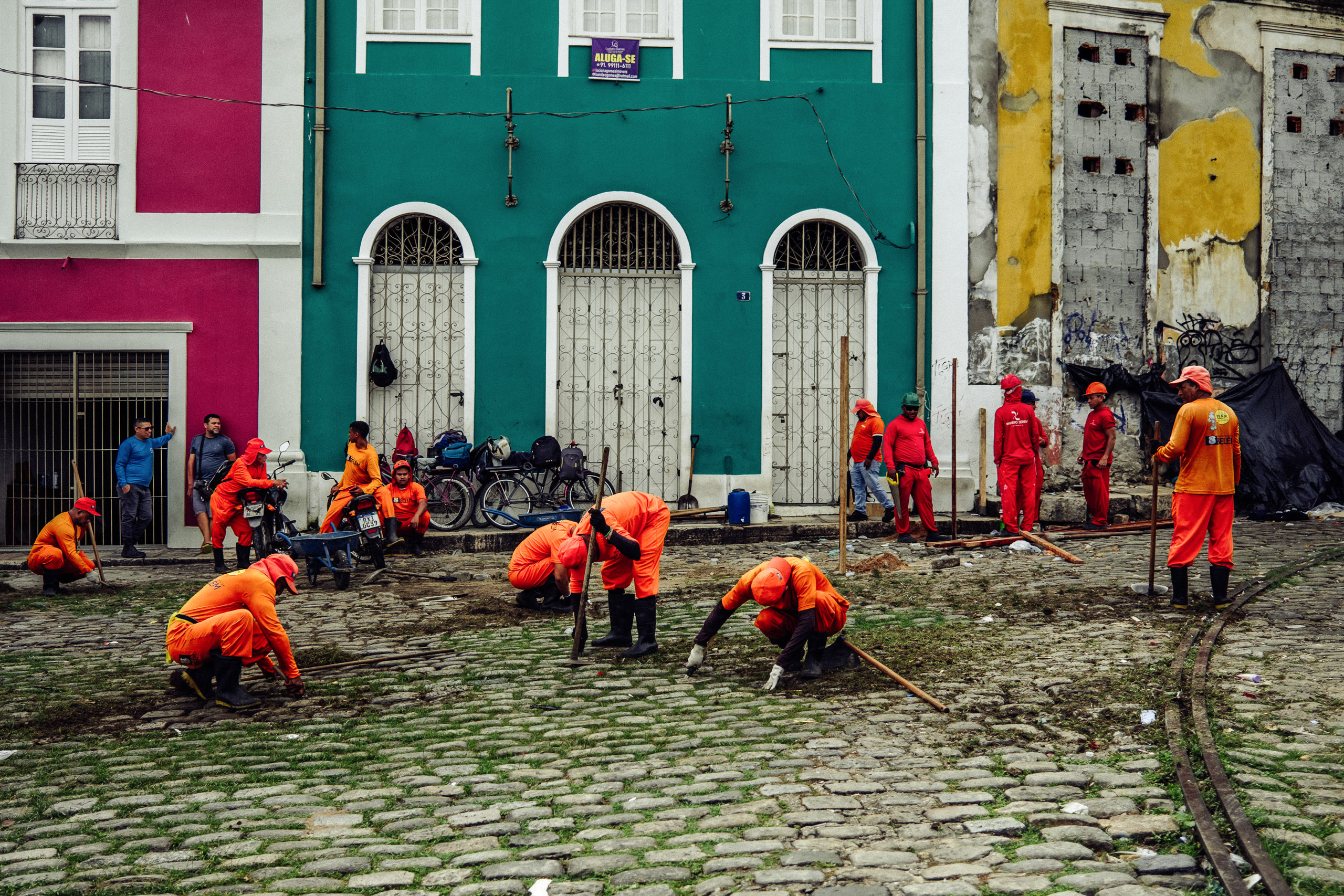 personas con uniformes naranjas quitando malas hierbas de una calle empedrada