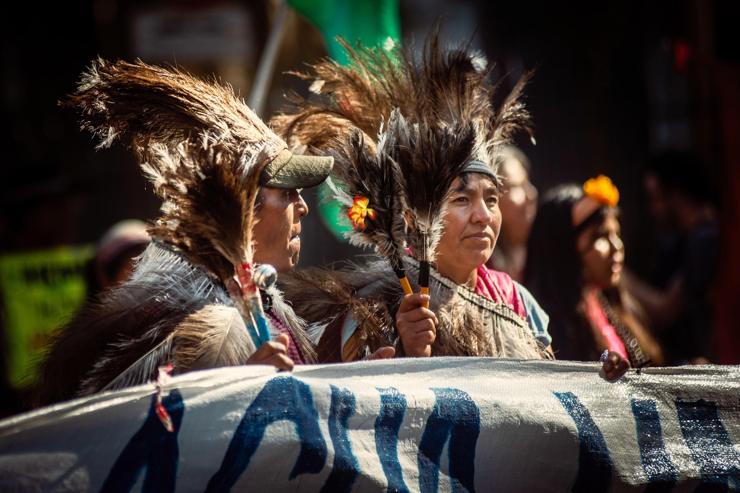 Dos manifestantes con plumas en la cabeza y una pancarta bajo la luz del sol