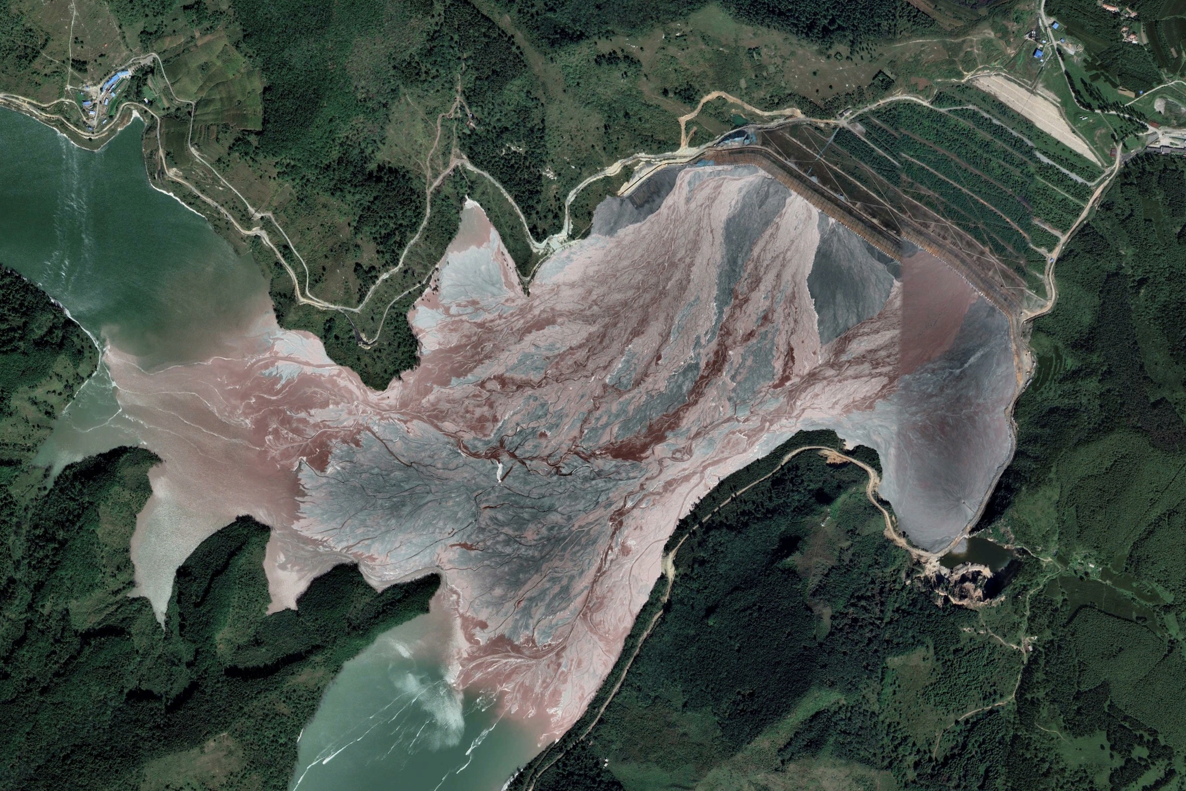 A satellite image of a vast reservoir with pink, red and blue stripes on the water, showing pollution from a mine