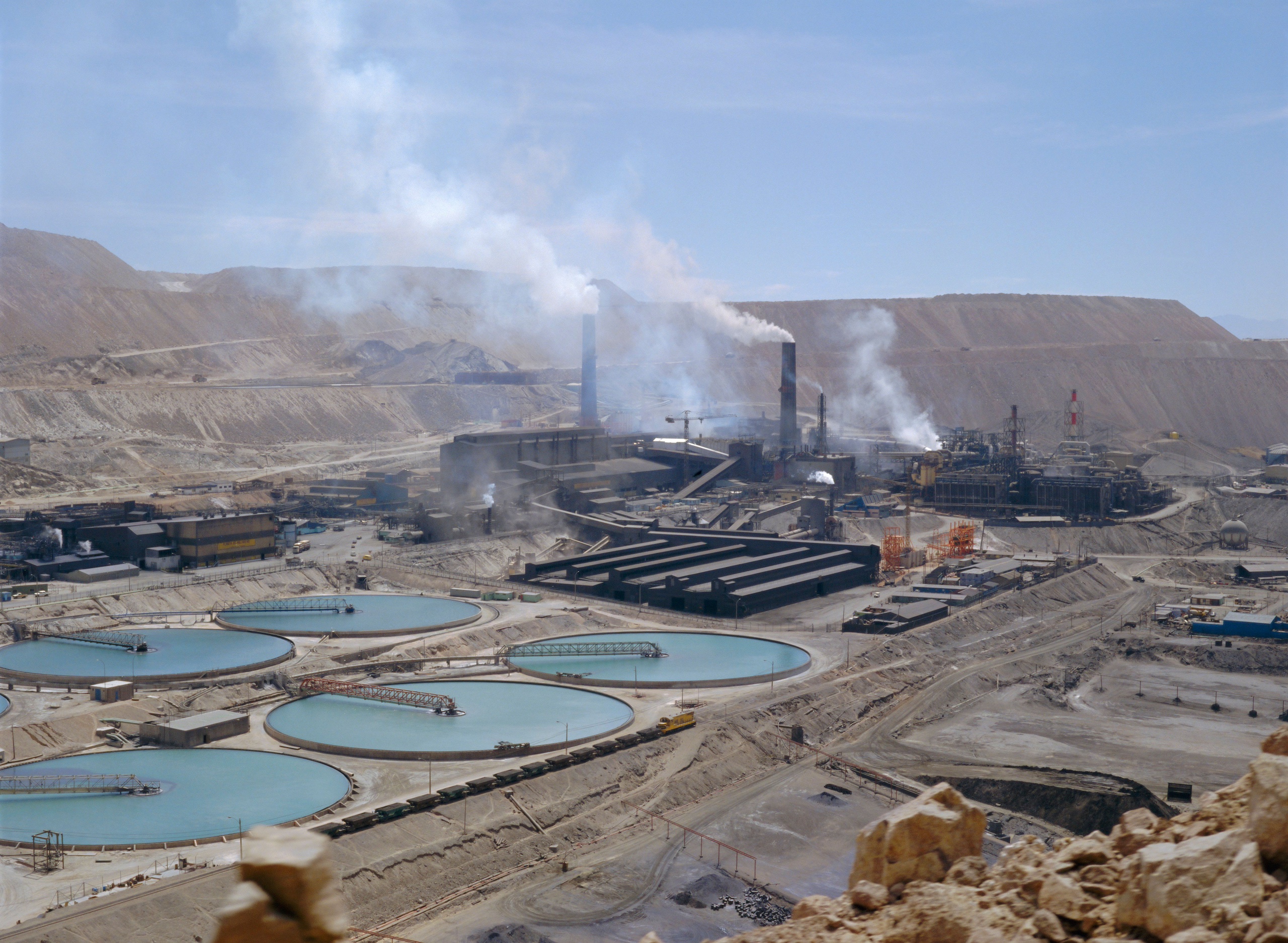 Industrial mining complex with circular water basins in the foreground, smokestacks emitting smoke in the background