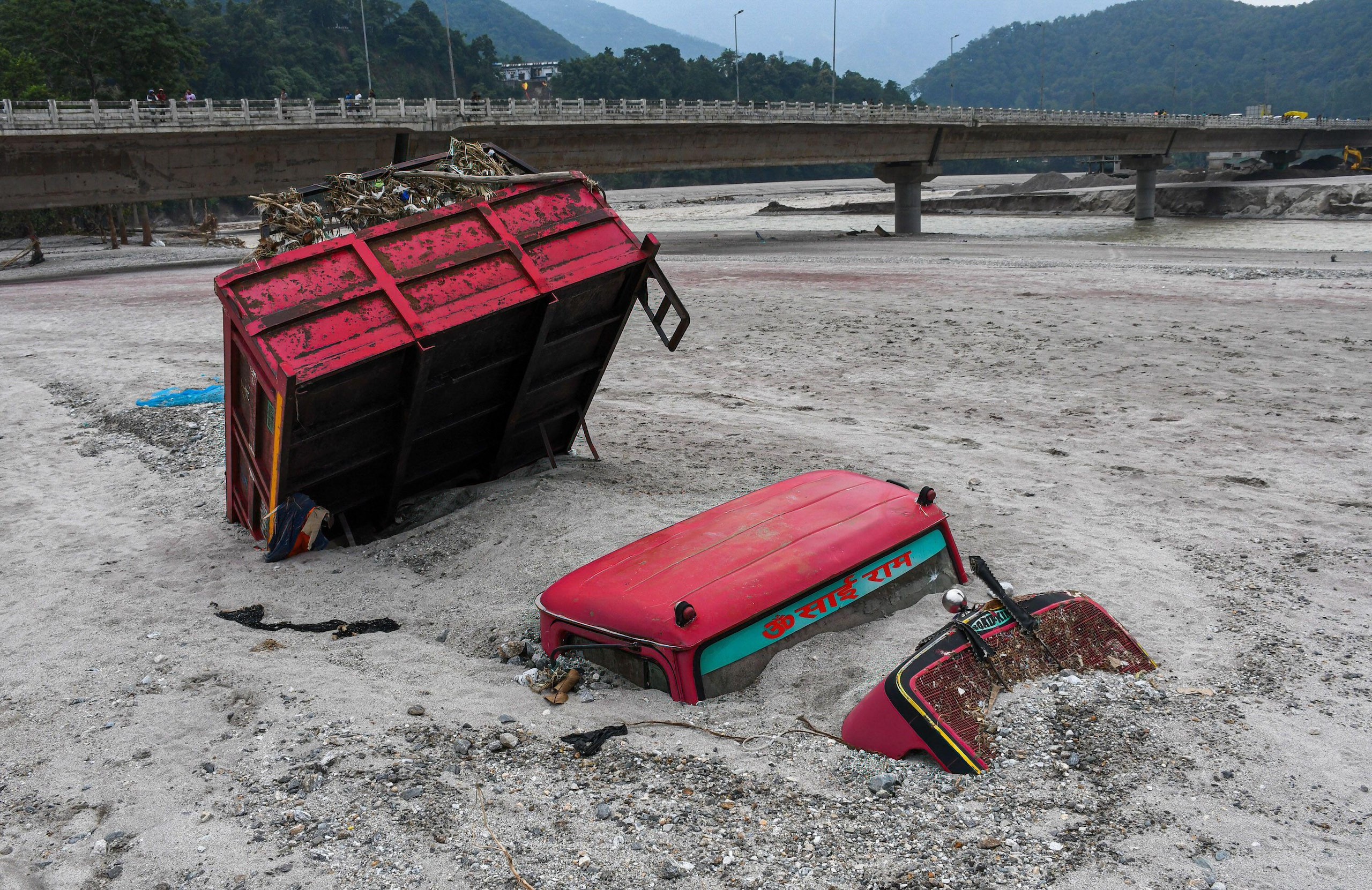 A red vehicle submerged in mud on a river bank, bridge in background
