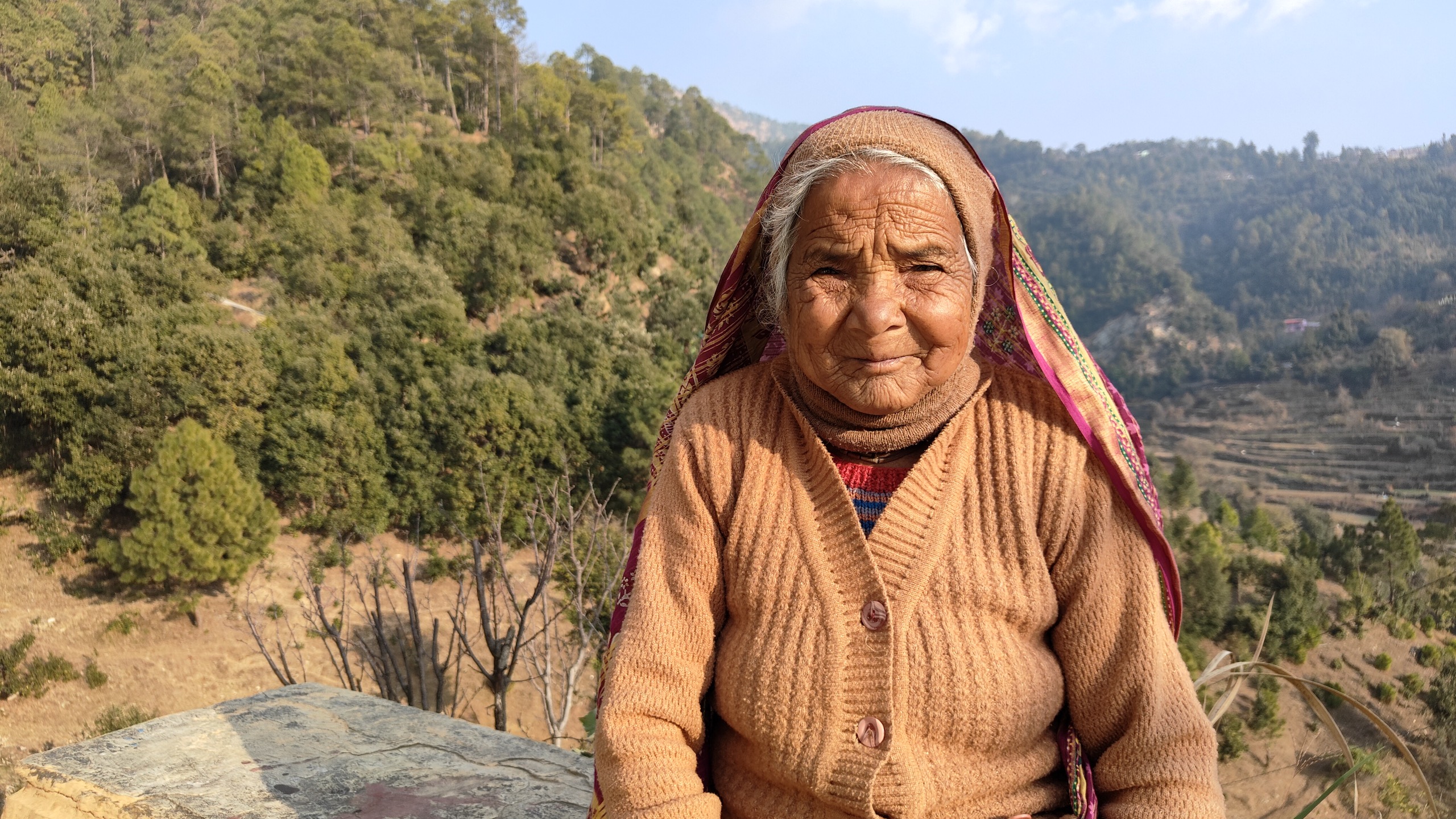 Elderly woman wearing a knitted sweater and a colorful headscarf, sitting in front of a scenic, forested mountain landscape