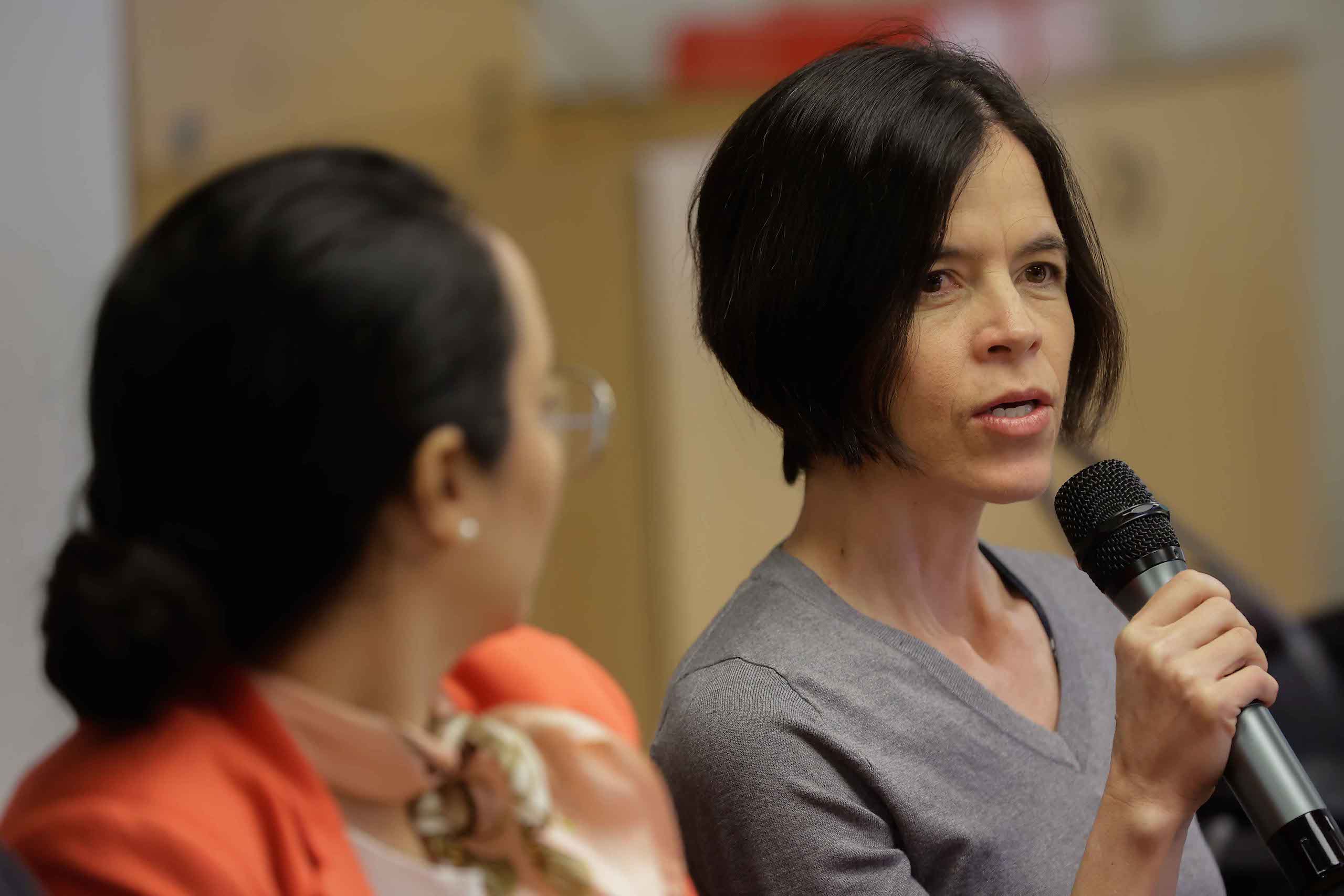 a woman addressing an audience through a microphone, with another woman observing