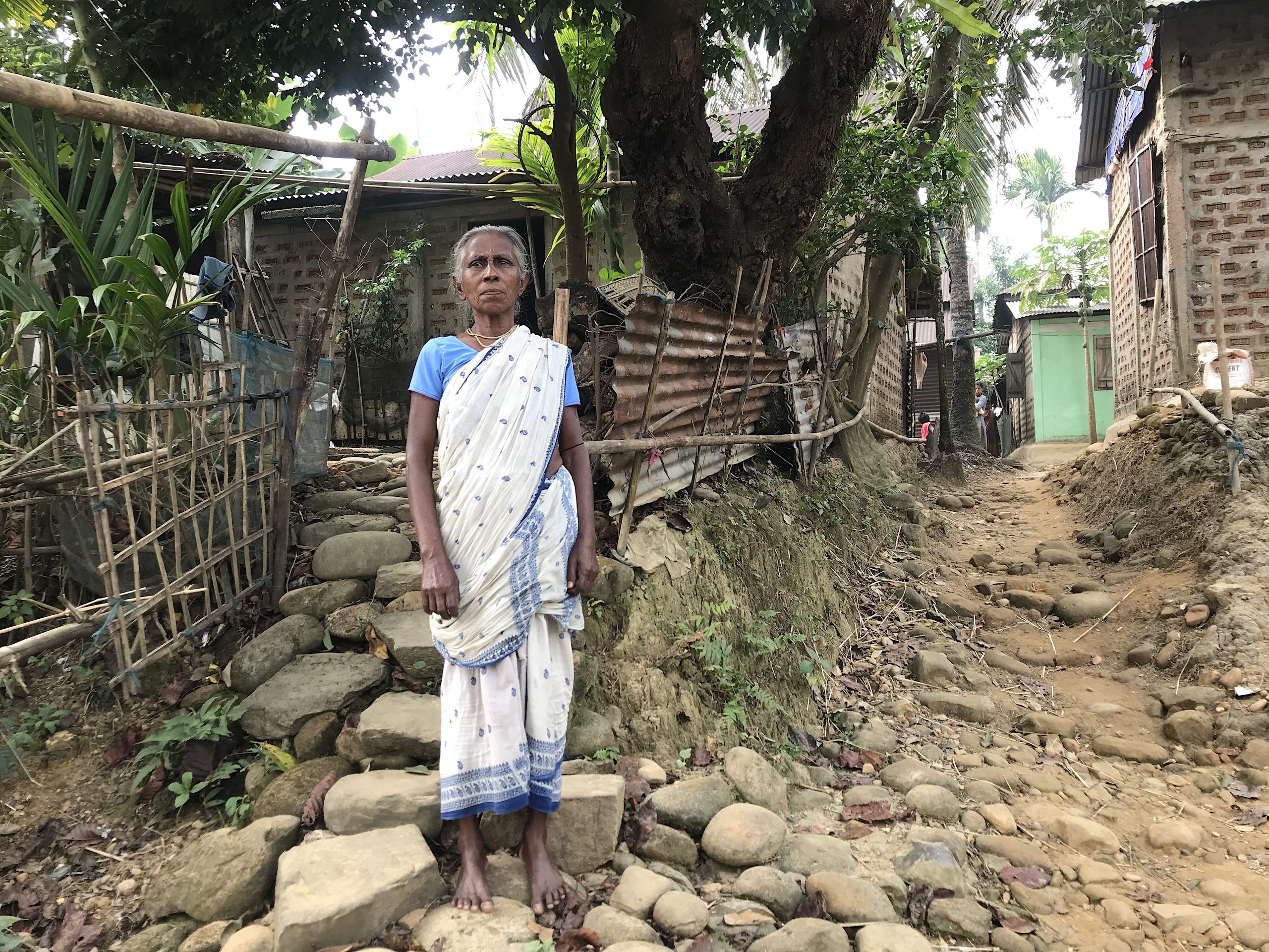 A woman stands on a dirt path in front of a house
