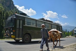 A man leading a horse looks at the camera, an army truck drives behind him along a mountain road