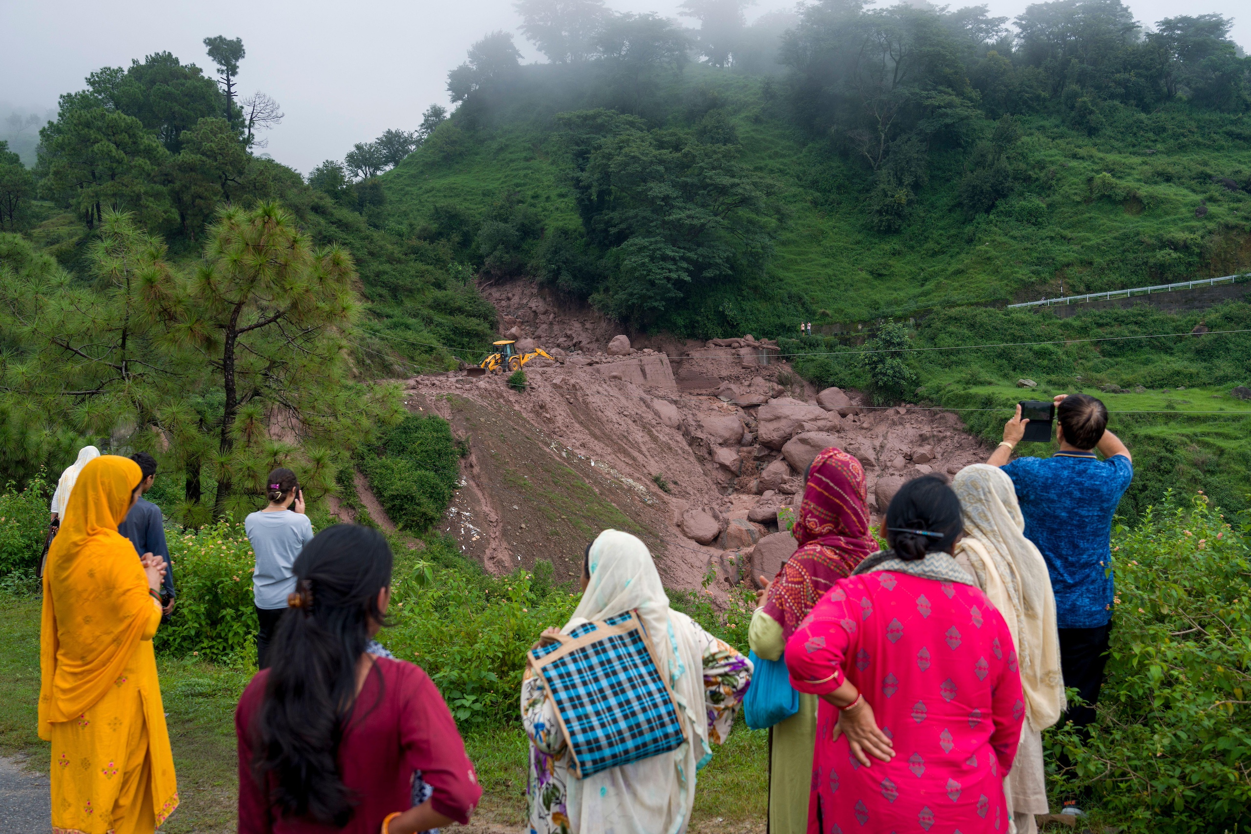 <p>People watch a digger moving debris after a landslide damaged a road near Dharamshala, August 14 2023. Heavy monsoon rains triggered floods and landslides in India&#8217;s Himalayan region, leaving several people dead and many others trapped. (Image: Ashwini Bhatia / AP via Alamy)</p>