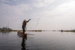 A fisher standing on a small boat on a calm lake casts a net