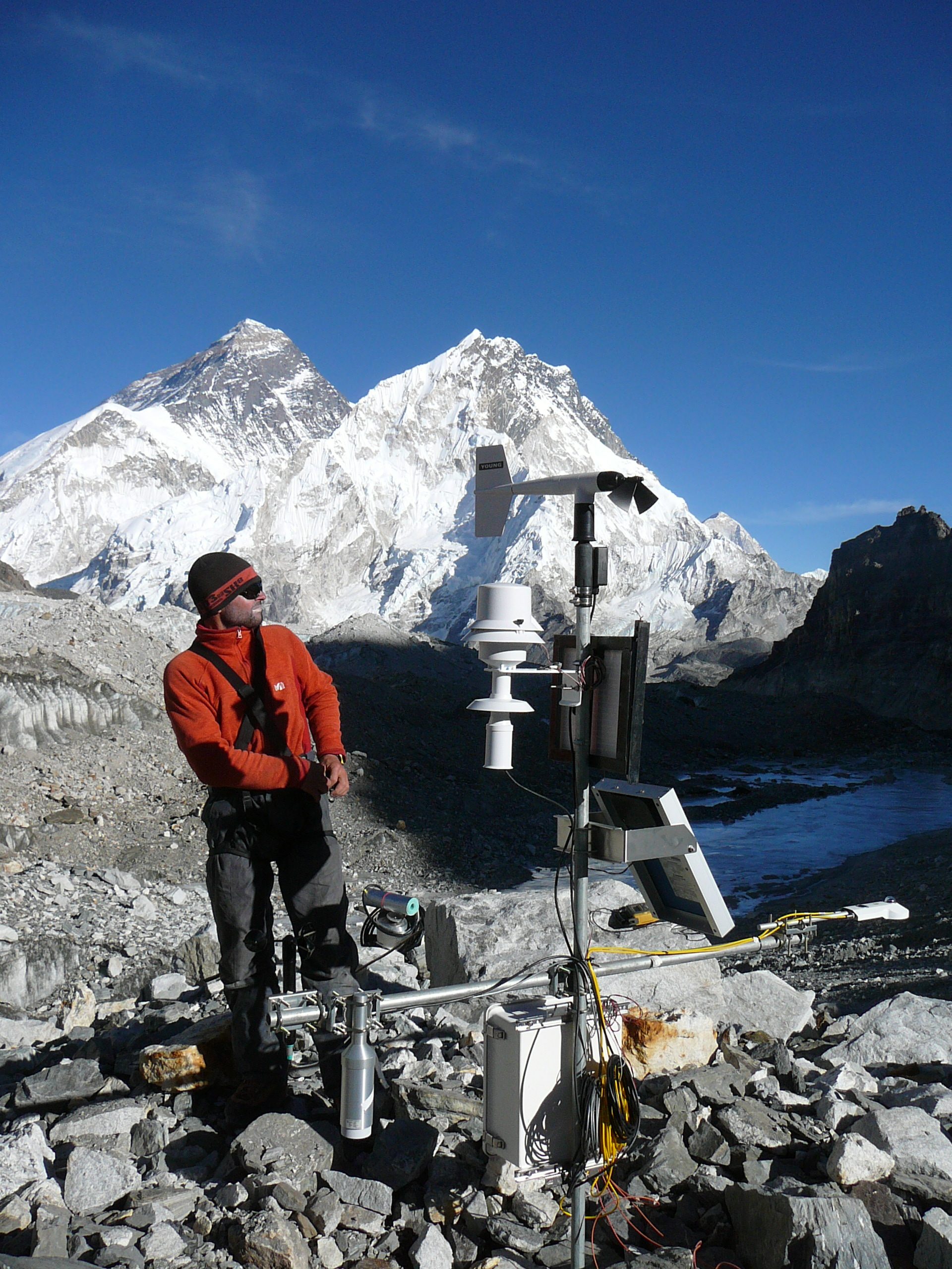 A man stands next to a weather station in front of tall snowy mountains