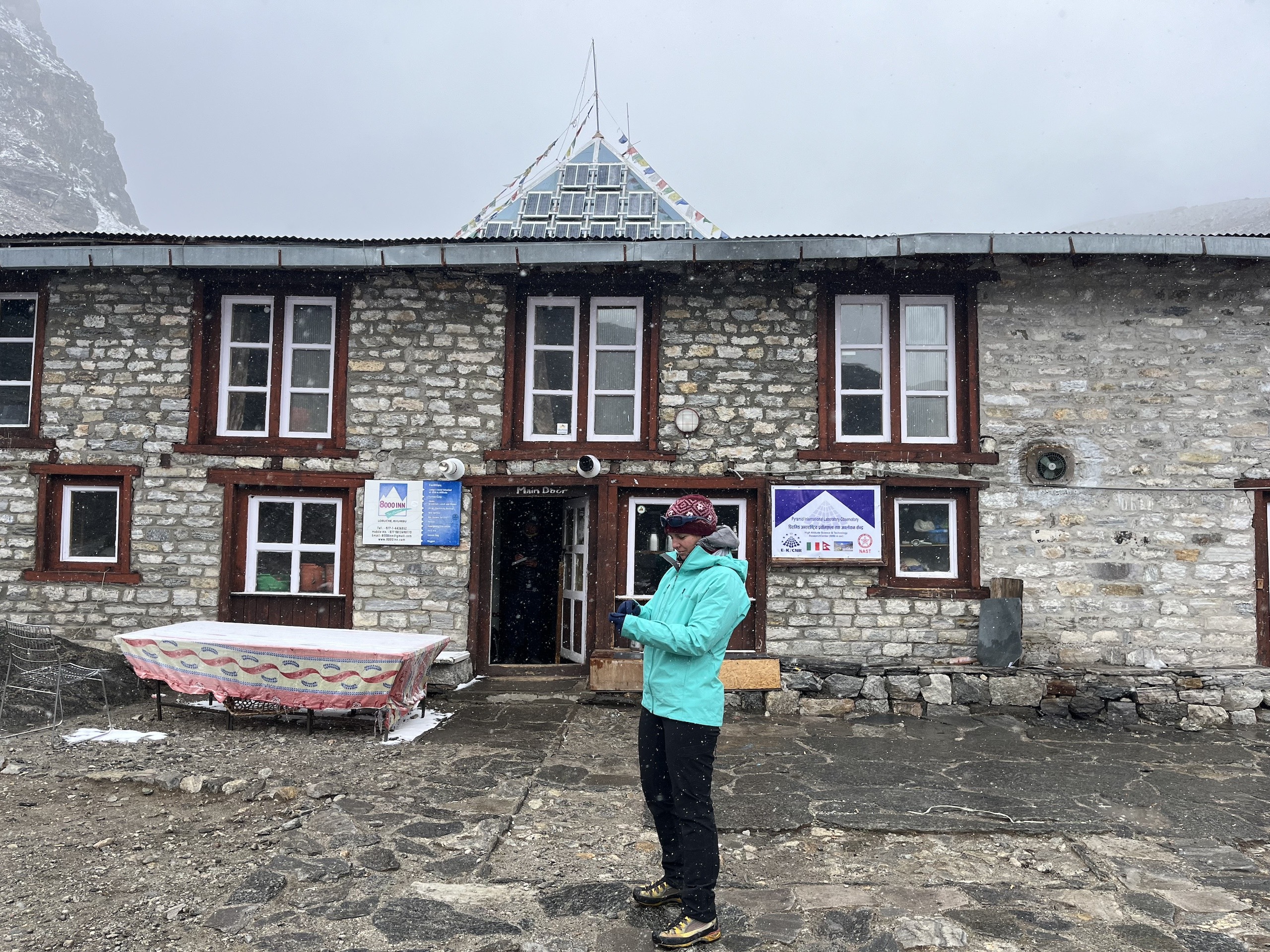 Person in a brightly coloured coat stands in front of a stone building in a misty mountainous area
