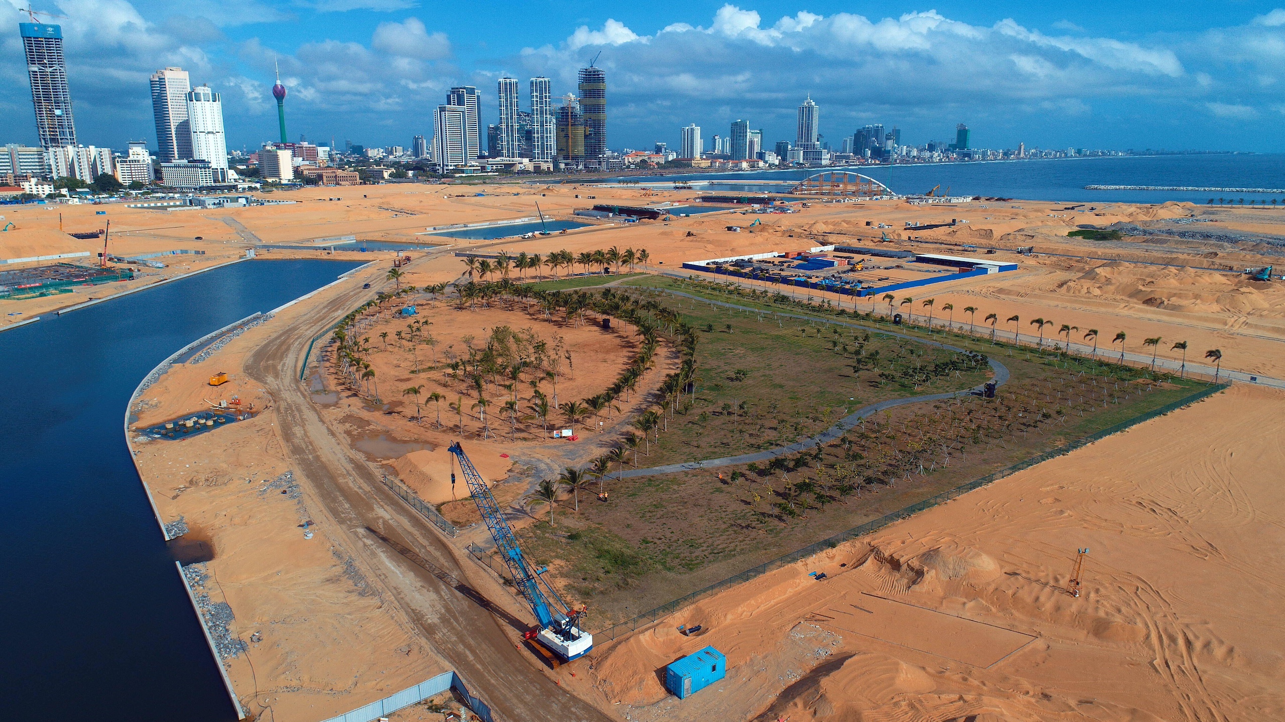 Aerial view of a large port under construction near a city, with a park lined with palm trees in the foreground