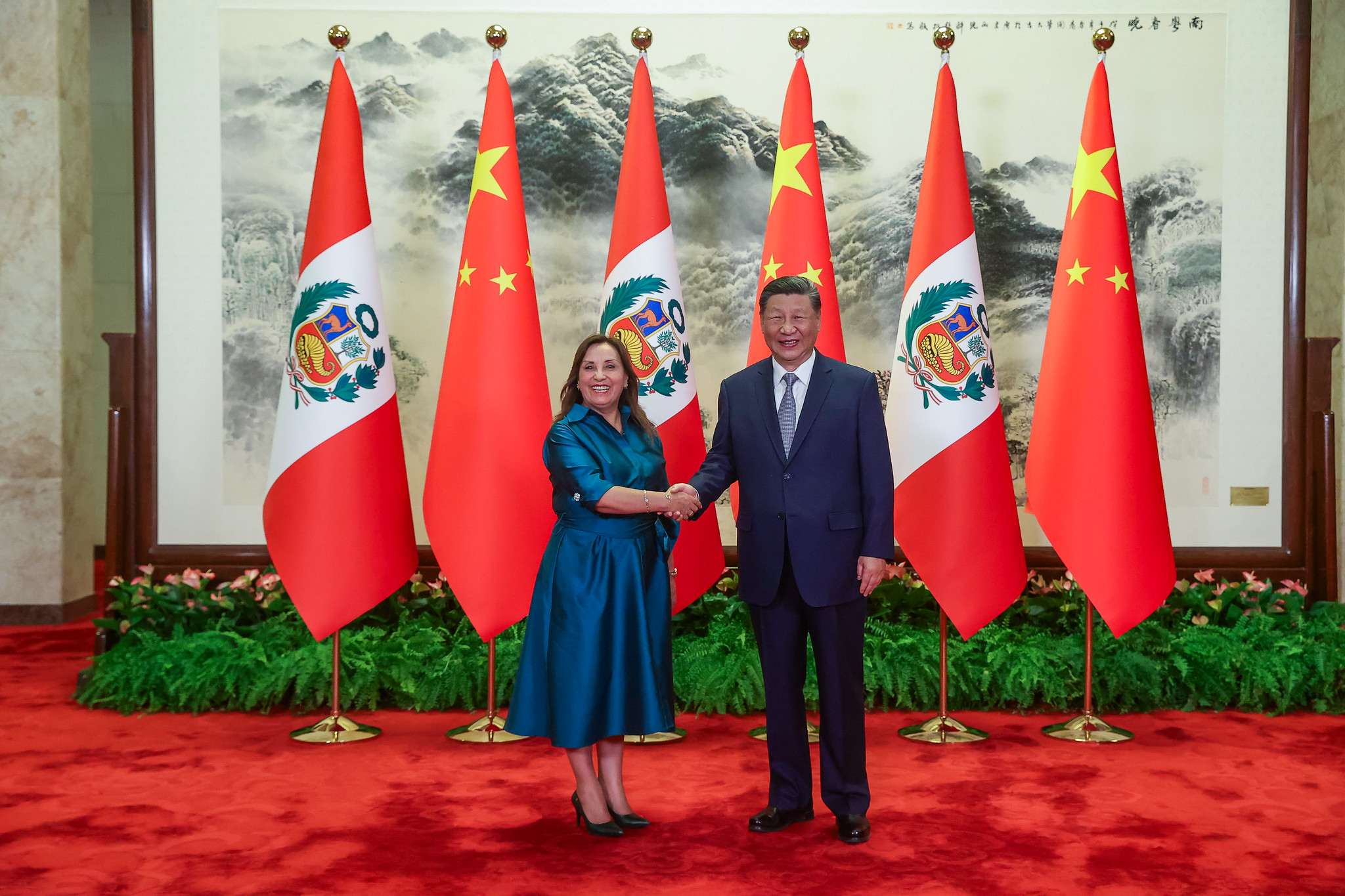 Dina Boluarte y Xi Jinping dándose la mano y sonriendo, parados frente a banderas de China y Perú