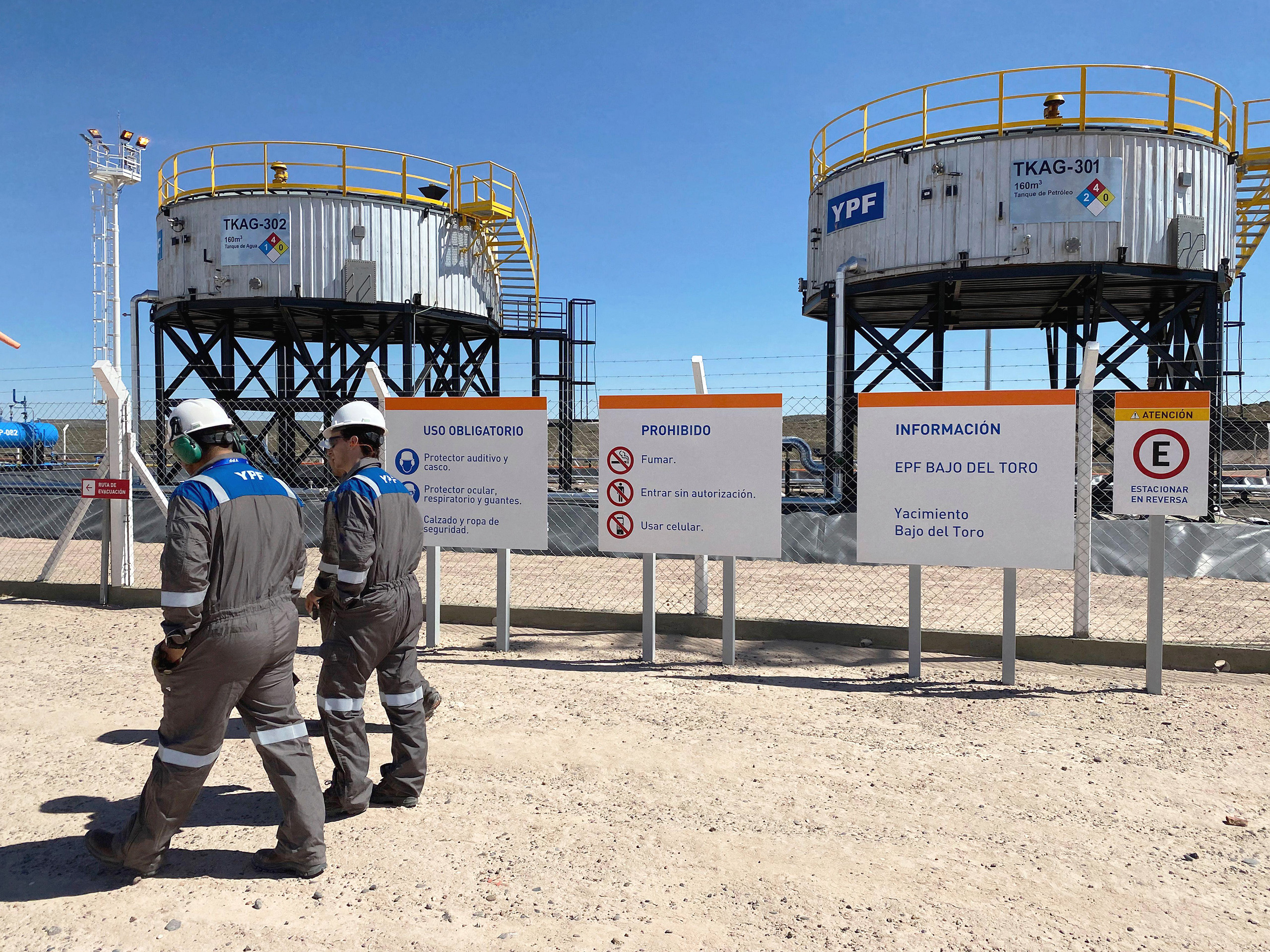 <p>Trabajadores de la petrolera estatal argentina YPF pasan frente a tanques de agua utilizados en la fracturación hidráulica en la cuenca de Vaca Muerta, provincia de Neuquén. El venteo, las fugas de metano y el uso de combustible son las principales fuentes de emisiones identificadas por la empresa en el yacimiento (Imagen: Titus Moser, Eye Ubiquitous / Alamy)</p>