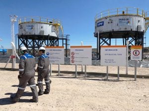 two men wearing protective gear walk in front of fenced oil tanks